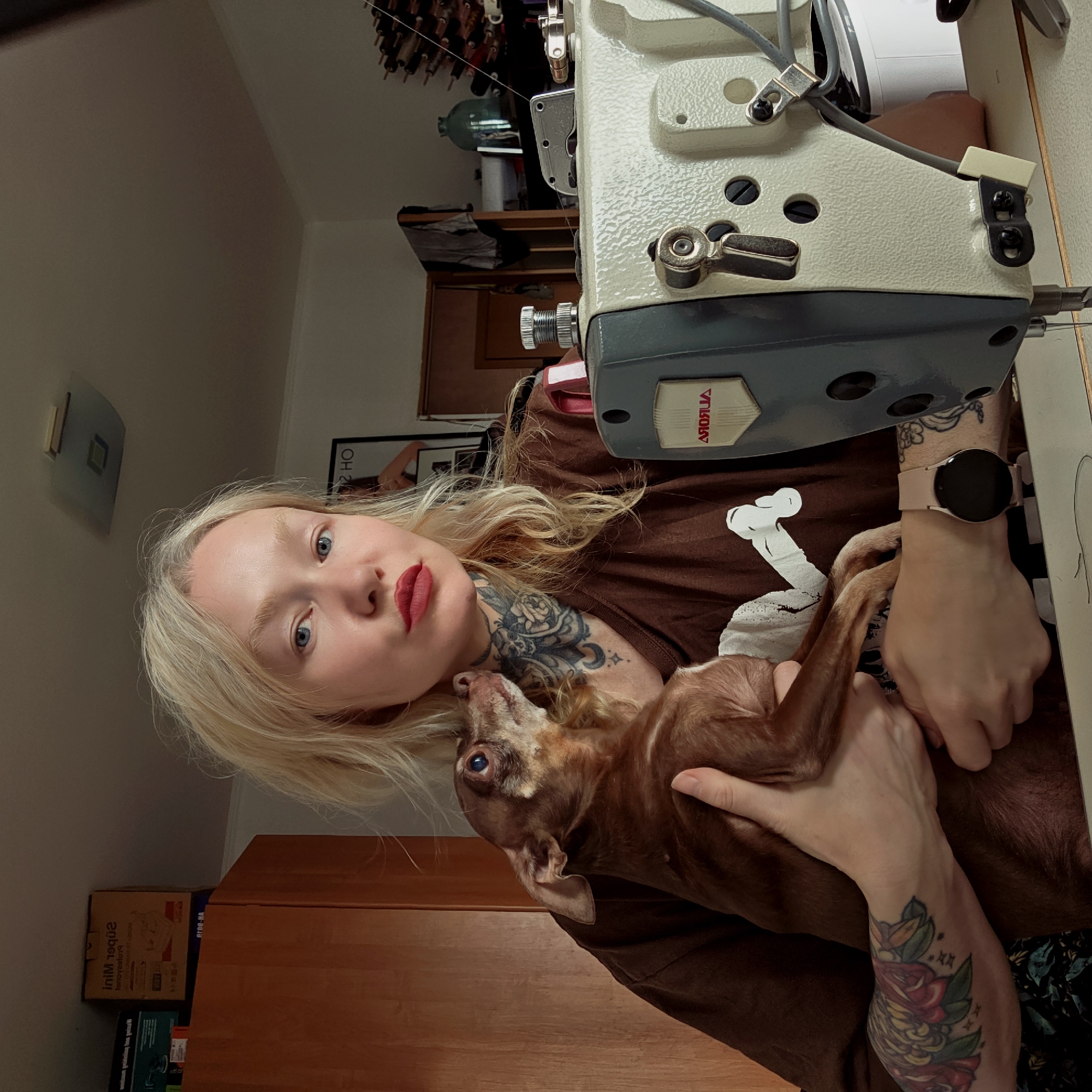 A woman with blonde hair and red lipstick is holding a brown Chihuahua in her arms. She is seated at a table next to a sewing machine. The setting appears to be a room inside a home. There is a sewing machine on the table in front of her. Tattoos are visible on her neck and arms. A pink watch is visible on her left wrist. Boxes and a cabinet can be seen in the background. Pictures are visible on the wall. Based on the sewing machine's nameplate (Aurora) and considering the request for the city of Saint Petersburg, Russia, it can be assumed the scene is taking place in that city.
