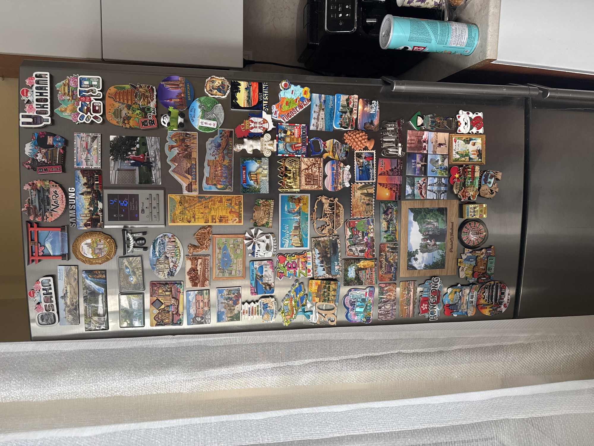 The image shows a stainless steel refrigerator door covered in souvenir magnets from various cities and countries. Text visible on the magnets includes "YOKOHAMA", "SHANGHAI", "PALESTINE", "EGYPT", "CRETE", "VIENNA", "SINGAPORE", "HONGKONG", "Dubai", "GREECE", and more. A magnet mentions "Василена и Веселин 01.09.2012", indicating an event for Vasilena and Veselin on that date. A Pringles can sits on top of the refrigerator, to the right of a black appliance. The lower portion of the image includes a white curtain or draped fabric. The location is not provided, and there is no mention of Plovdiv, Bulgaria.