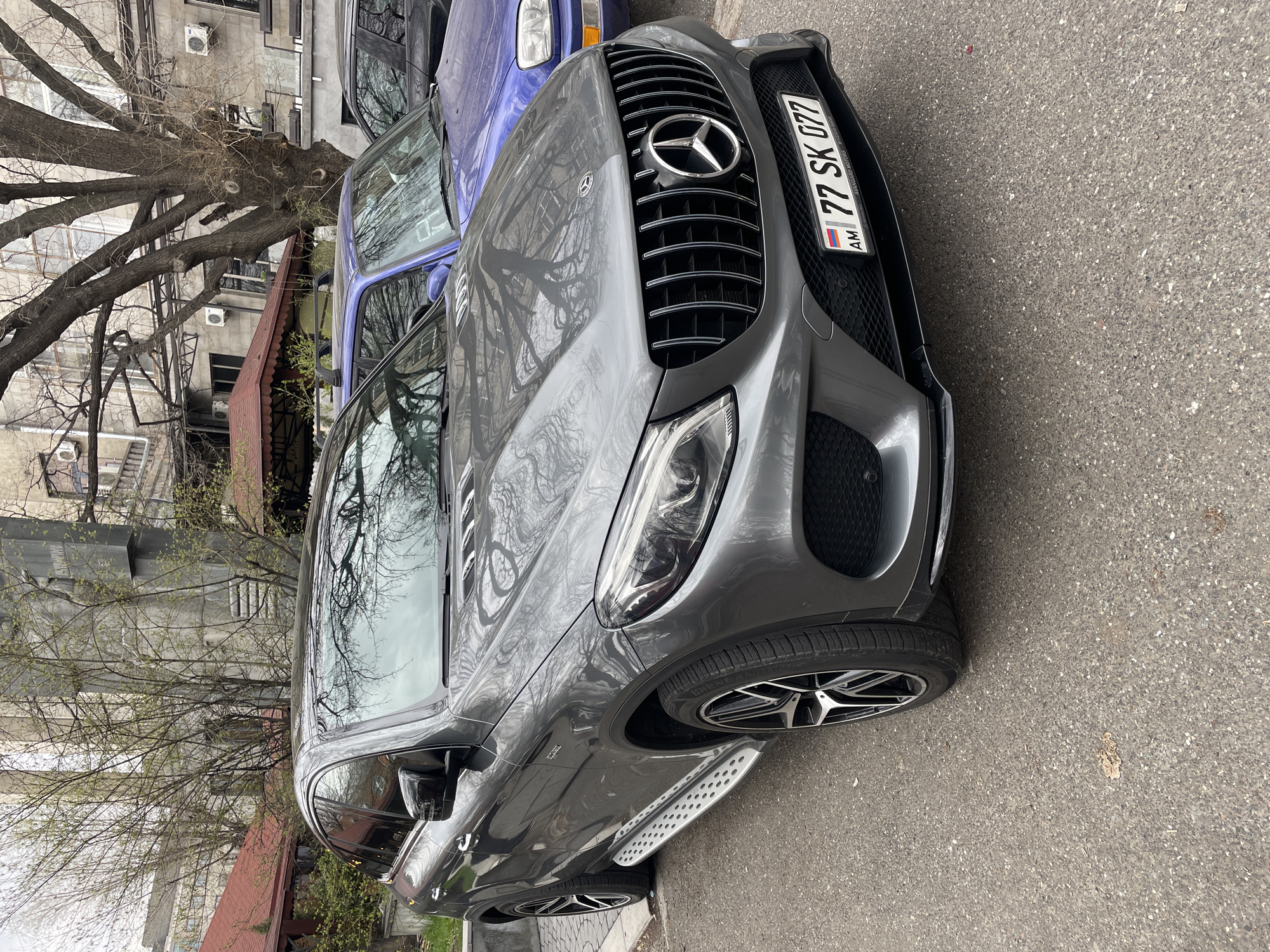 A gray Mercedes-Benz SUV is parked on a Yerevan, Armenia street. The car's license plate number is "77 SK 077". A blue car is parked behind it. A tree and buildings are in the background.
