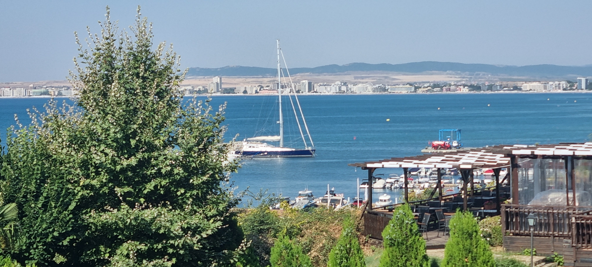 The image depicts a scenic view of a coastal city with a body of water in the foreground and a distant cityscape. The water is a deep blue and calm, with a sailboat visible in the middle ground. The sailboat is a large, white vessel with tall masts. Along the shoreline of the distant city, there are numerous buildings, including hotels and apartments, and a Ferris wheel can be seen in the cityscape.
In the immediate foreground, there is a lush green tree with small white flowers, obscuring a portion of the view of the water and the city. To the right of the image, there is a structure that appears to be an outdoor dining area with wooden tables and chairs, and a covered roof. Beyond this dining area, a marina is visible, filled with numerous smaller boats docked in the water. In the far right of the image, on a pier extending into the water, there is a blue structure that resembles a boat lift or a crane, with a red object next to it, possibly a vehicle. The sky is clear and blue, suggesting a sunny day.
The overall scene is one of a pleasant coastal resort town. Based on the architectural style of the buildings and the presence of a marina, the location is likely a tourist destination. The image captures a typical summer day at a seaside resort, with a mix of natural beauty and man-made structures.