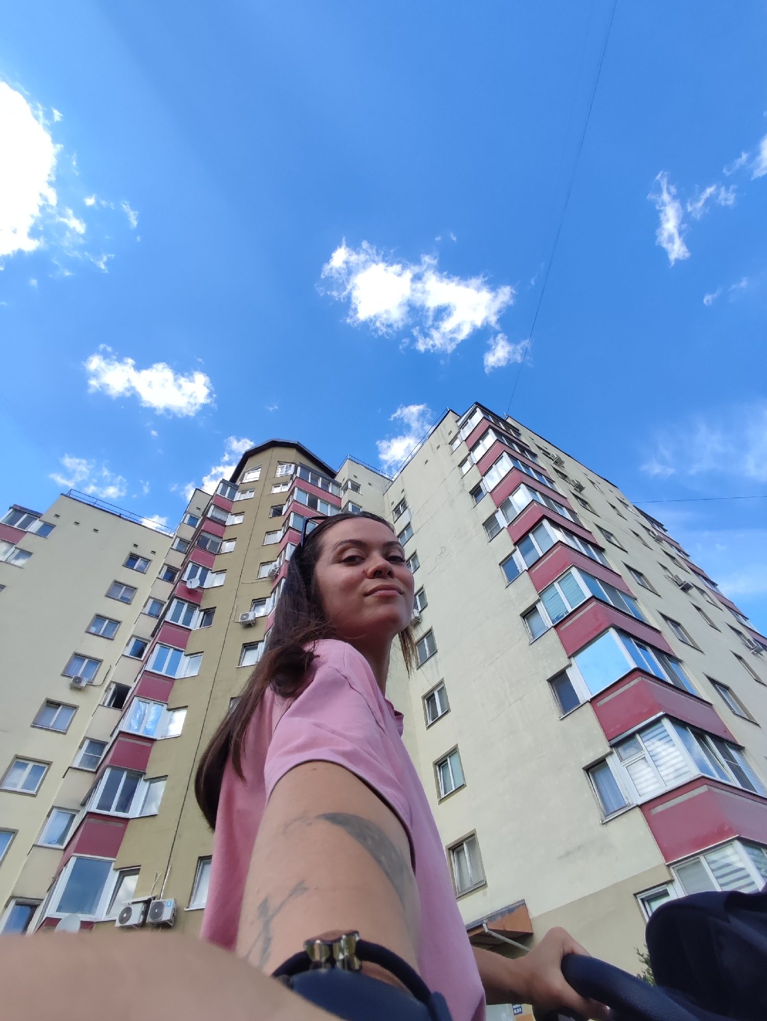 The image is a low-angle, selfie-style photograph taken outdoors, likely in the city of Chaikovsky, Russia. The foreground features a young woman with long, dark hair, wearing a pink t-shirt. She is looking towards the camera with a slight smile. Her right arm and part of her torso are visible, and she has tattoos on her arm. She appears to be holding onto the handle of a stroller, which is partially visible in the lower right corner. In the background, a tall, multi-story apartment building dominates the scene. The building is painted in light yellow and has accents of red around the balconies and windows. The windows are numerous and varied in their appearance, with some reflecting the blue sky. The sky itself is a clear, bright blue with scattered white clouds. A thin black wire is visible stretching across the upper portion of the sky. The perspective suggests the photograph was taken on a sunny day.