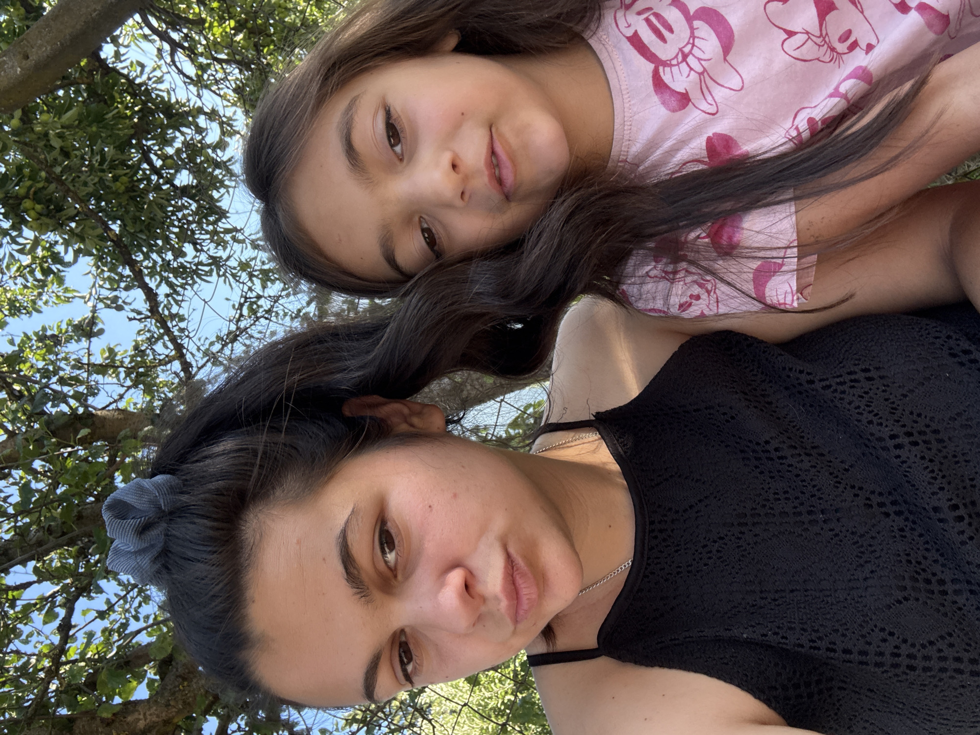 The media file is a selfie taken outdoors, featuring a woman and a young girl positioned closely together. The woman, in the foreground, has dark hair pulled back with a grey scrunchie and is wearing a black, lacy top with a silver necklace. The young girl, positioned behind and above the woman, has long dark hair and is wearing a pink t-shirt with a repeating Minnie Mouse pattern.
The setting appears to be a natural environment with trees and blue sky visible in the background. The lighting suggests it is daytime, likely sunny. The woman's expression is relatively neutral, while the girl is smiling. The image is oriented vertically, with the subjects tilted. There is no visible text. The location context is Oreshak, Bulgaria.
