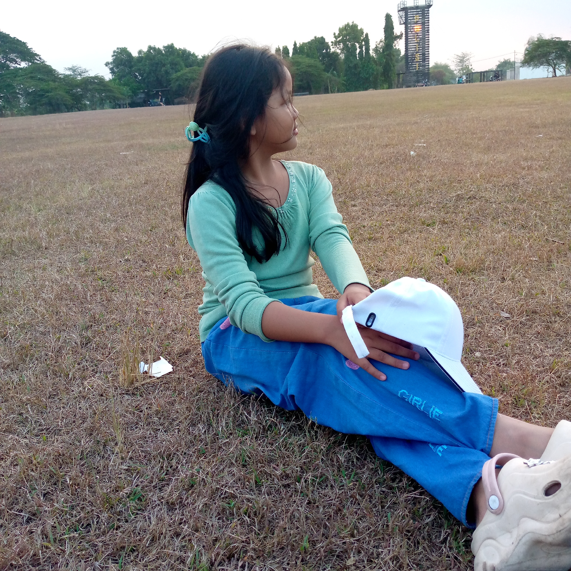 A young girl with long, dark hair pulled back with a blue hair tie sits on dry, brown grass in an outdoor area, likely a park or field, in Pacing, Indonesia. She is wearing a light green long-sleeved shirt with beaded embellishments around the neckline and bright blue jeans. She is looking to her right, away from the camera. In her hands, she holds a white baseball cap. Her feet are clad in light-colored slip-on shoes with pink straps. The background shows a sparse collection of trees, a tall, dark structure resembling a tower with lights, and some motorcycles parked in the distance. The ground is covered in dry, matted grass with a few scattered pieces of white debris. The sky is overcast.