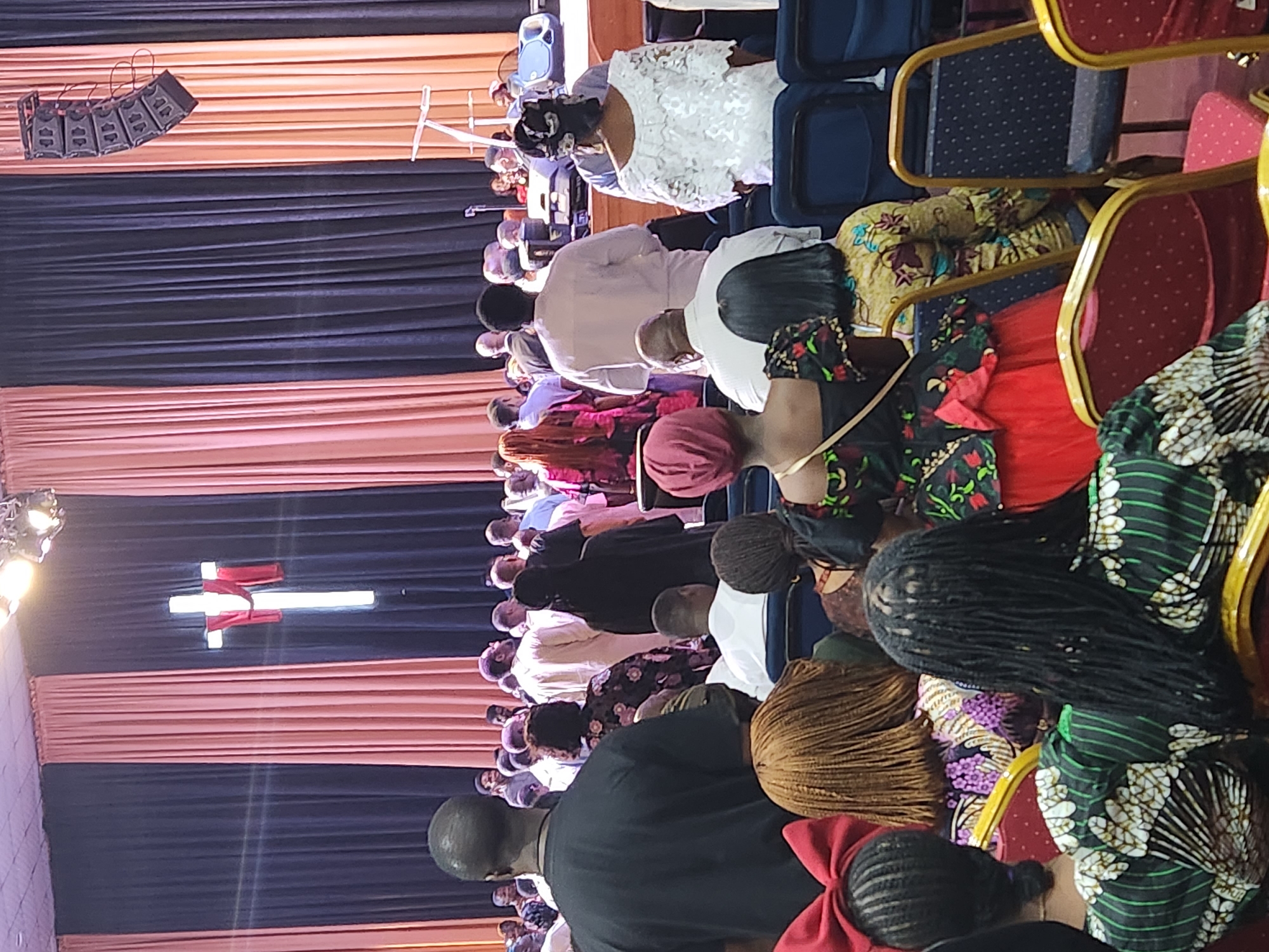 In a large indoor venue, likely a church in Barakin Lerawi, Nigeria, a congregation is gathered. The background features a stage area with dark and peach-colored drapery. A prominent illuminated cross with a red sash hangs on the stage, with a large speaker system suspended from the ceiling to the right. In the foreground, the backs of several congregants are visible, seated on chairs. They are dressed in vibrant, patterned attire. The overall atmosphere appears to be one of a religious service or gathering.