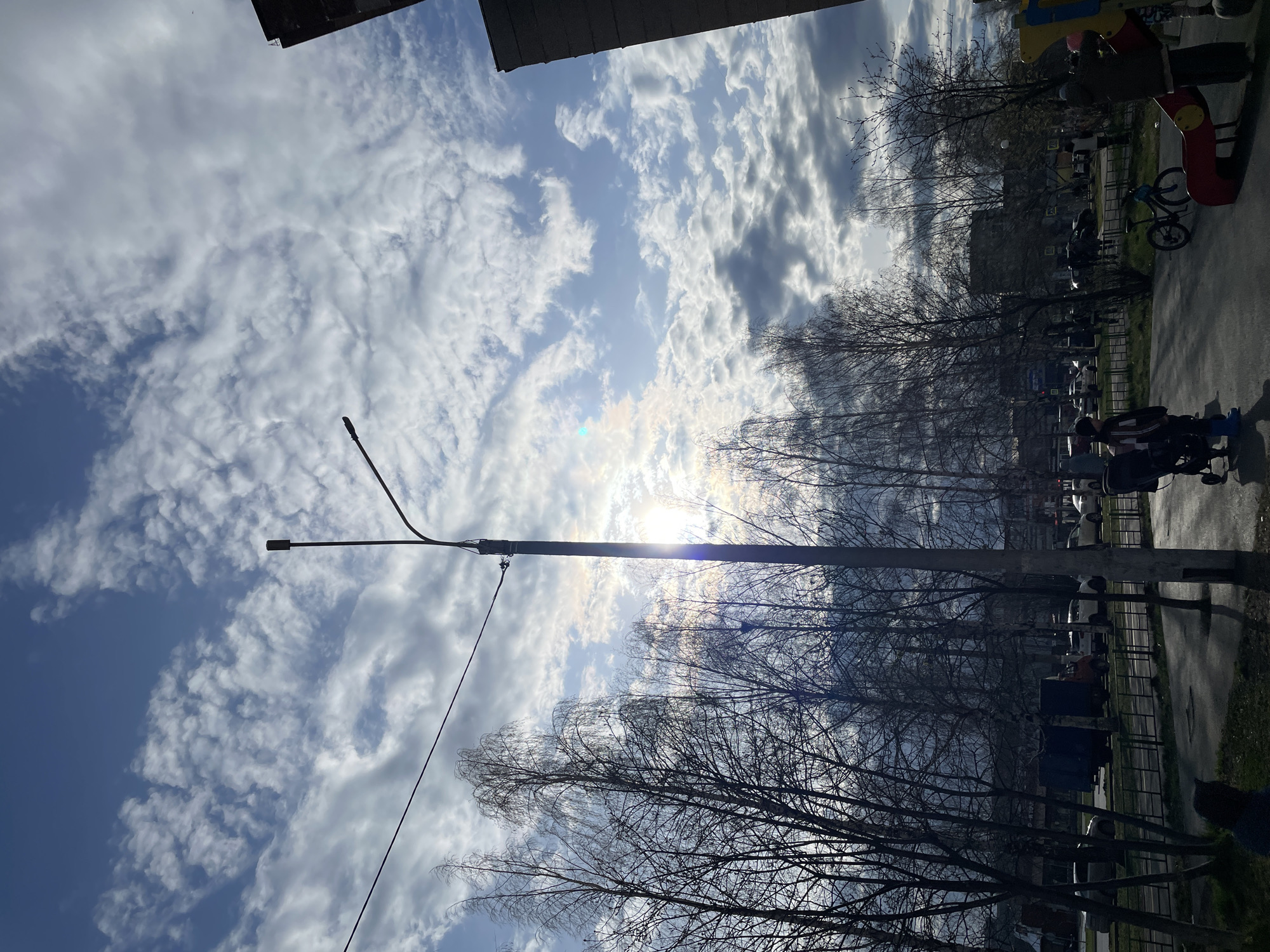 The image shows a view in Izhevsk, Russia. The scene captures a streetscape with a mix of natural and urban elements. A tall, metal lamppost dominates the foreground, extending from the bottom of the frame towards a bright, partially obscured sun, which is surrounded by a blue sky filled with white, patchy clouds. Silhouetted tree branches reach across the middle of the frame, obscuring parts of buildings. On the right side of the image, a paved area, a road, is visible with a section of green grass. The image includes several people and a bicycle near a playground. The architectural elements include the tops of buildings, some with visible roofing. There appears to be light traffic on the road.