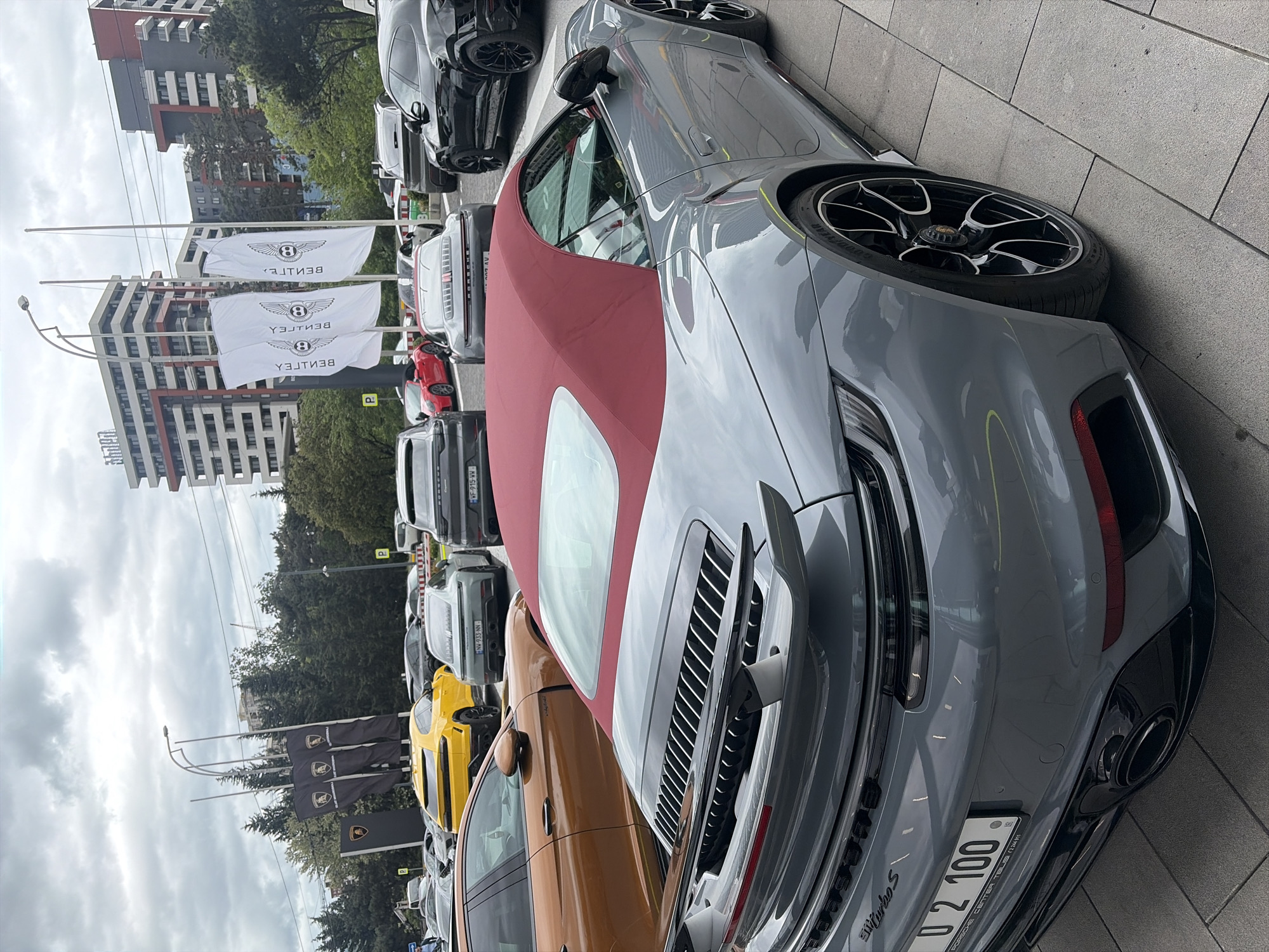 Here is a detailed description of the image:
**Scene Overview:** The image shows a lineup of luxury cars parked along a paved area, seemingly adjacent to a dealership or high-end car exhibition. The location can be inferred to be in Tbilisi, Georgia, based on the visible license plates and the architectural style of the buildings in the background.
**Objects and Subjects:**
* **Cars:** The main focus is on several high-end vehicles. A gray Porsche 911 Turbo S convertible with a red top is prominently featured, taking up a significant portion of the frame. Other cars include what appears to be Bentleys and Lamborghinis, based on the flags and some visible vehicle designs.
* **Buildings:** Multi-story modern buildings stand in the background, indicating an urban setting.
* **Flags:** Bentley flags are visible, as well as Lamborghini flags.
**Environment:**
The setting is urban, with modern buildings and what seems to be a well-maintained paved area.
**Conclusion:** The scene depicts a luxury car showcase in an urban environment, likely near a dealership in Tbilisi, Georgia.