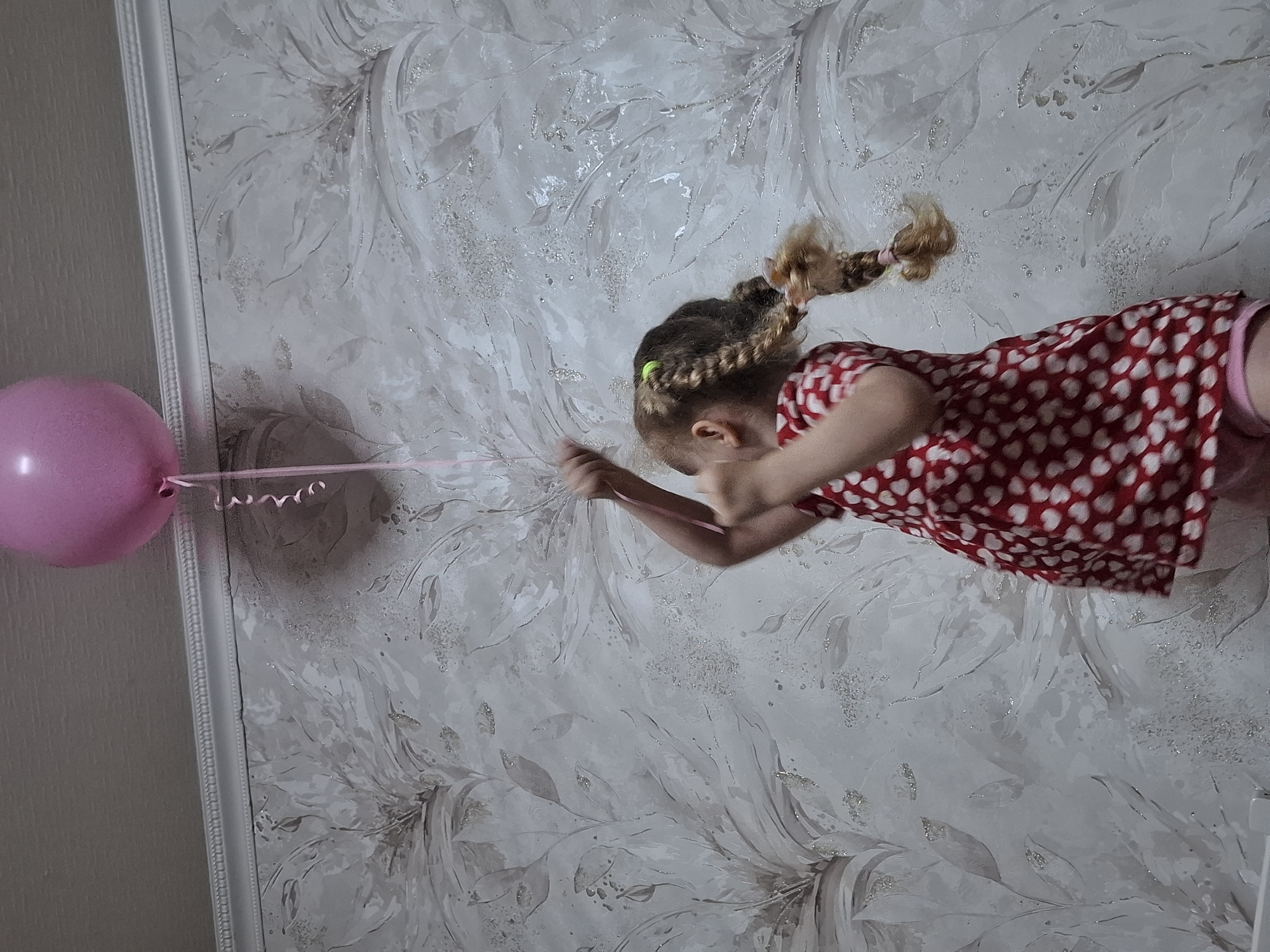 A girl with blonde hair braided into pigtails, wearing a red and white heart-patterned dress, is pulling on the string of a pink balloon. The girl is positioned against a wall with a floral wallpaper pattern. The location is Chaikovsky, Russia.