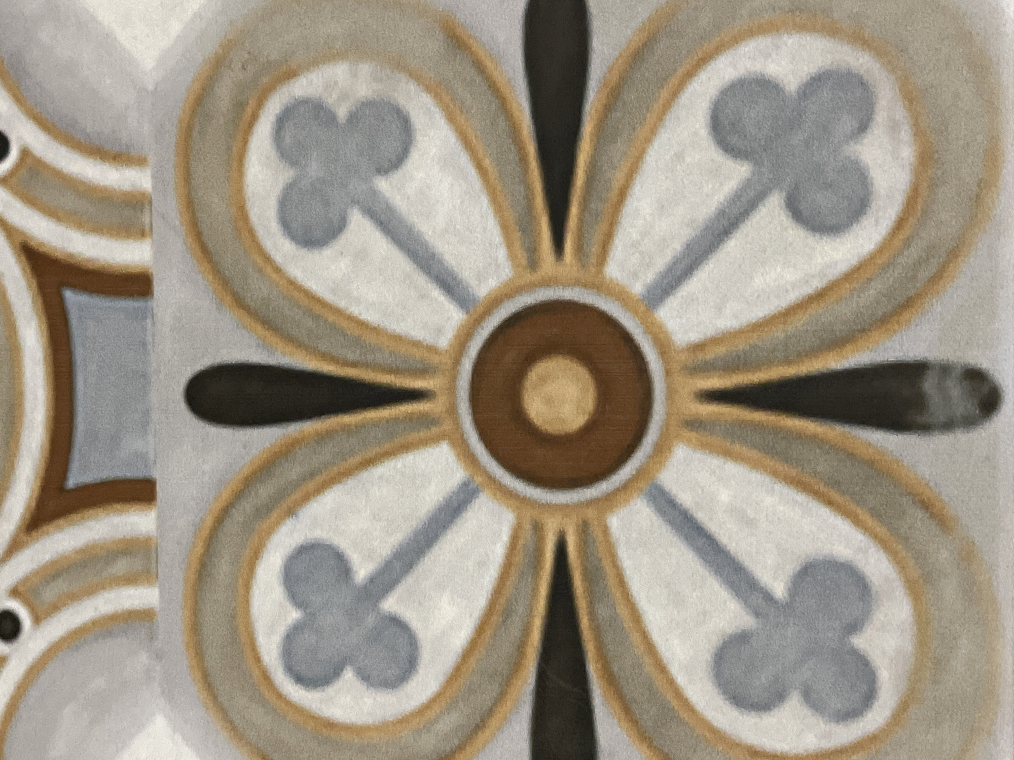 The image is a close-up shot of a decorative tile pattern, likely part of an architectural element in Tbilisi, Georgia. The pattern features a central circular motif with four outward-radiating petal-like shapes, each containing a stylized clover-like design in a muted blue-grey color. These shapes are outlined in a thin gold band and then a wider grey-brown band. The entire design is set against a textured off-white background. The top of the image shows a sliver of another decorative element, with curved bands of white, gold, and brown, and a small black circle. The lighting is even, and the image appears to be taken at a slight distance from the surface, emphasizing the intricate details of the tile work. There are no people or any dynamic actions depicted. The style suggests it could be found in an older building, possibly a church or a historical apartment block, common in Tbilisi.