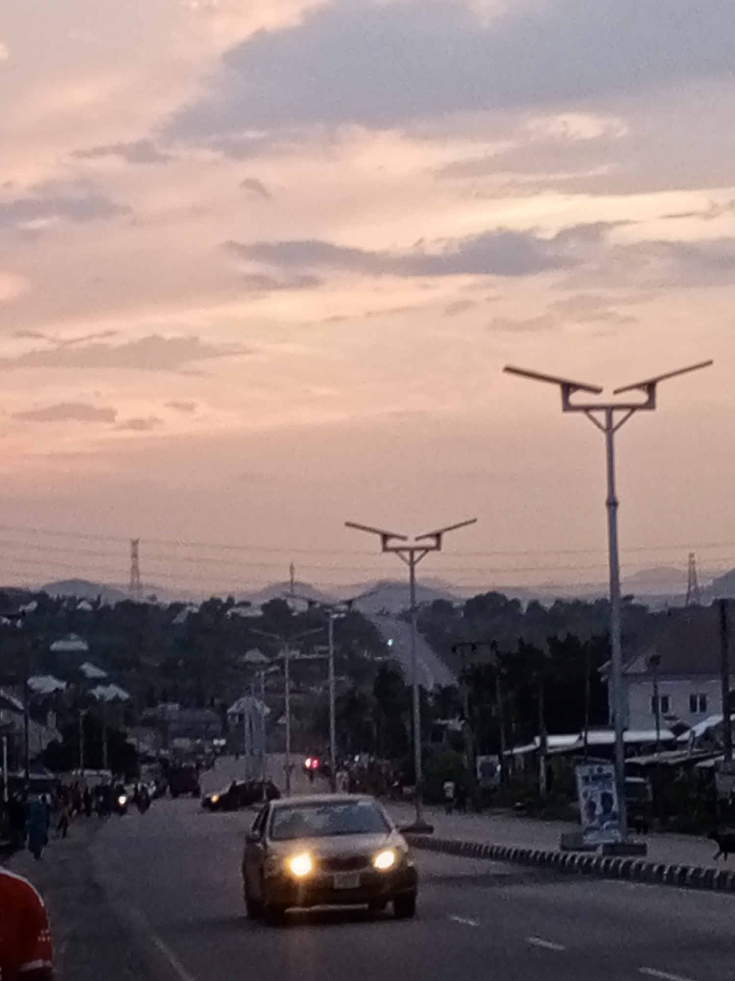 The image depicts a street scene at dusk in an unidentified city, possibly near a mountainous region, given the visible hills and transmission towers in the background. The sky is a mix of muted pink and grey. A car with headlights on is traveling on the road towards the camera. There are streetlights along the road. On either side of the street, there are buildings and pedestrian activity. In the background, other vehicles can be seen moving along the road.