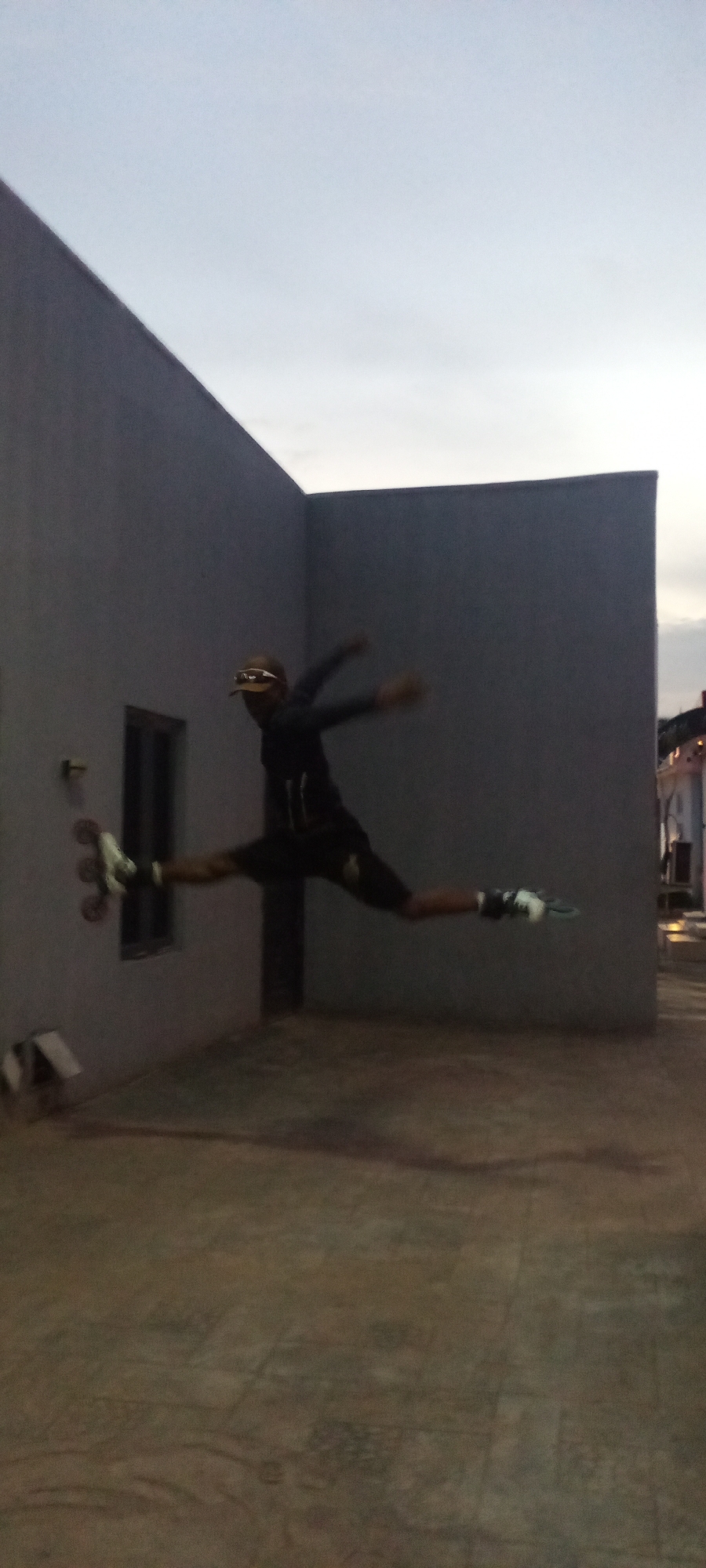 A dynamic outdoor shot captures a person mid-air in Akwanga, Nigeria, performing an athletic split jump while wearing rollerblades. The individual is dressed in dark attire, including a hoodie and shorts, and sports a cap or visor. Their body is horizontally extended, with legs splayed apart in a grand jeté-like pose, showcasing agility and balance on the rollerblades which are clearly visible on their feet.
The scene is set in an enclosed outdoor space, possibly a courtyard or rooftop, defined by two plain, light grey walls forming a corner. A small, dark rectangular window is visible on the left wall. The ground beneath is a dark, rough-textured surface, likely concrete or pavement.
The lighting suggests twilight—either early morning dawn or late evening dusk. The sky above is a soft, muted grey-blue, indicating low ambient light conditions. The overall atmosphere is dim, contributing to the focus on the central figure's powerful movement. There is no visible text in the image.