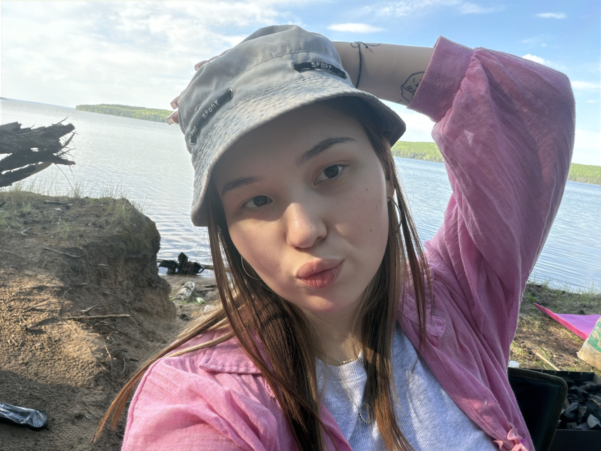 A young woman is taking a selfie outdoors with a lake in the background. She is wearing a grey bucket hat with the word "SPORT" on it, a pink long-sleeved shirt, and a grey t-shirt. She is holding her hat with her left hand. There is a tattoo on her left arm. The environment includes a grassy area with a sandy bank, and a body of water, likely a lake, in the distance. Some tree-covered islands are visible on the other side of the lake under a blue sky with some clouds. There is also a charcoal grill with charcoal bricks in it and a triangular pink tent peg in the background. The location appears to be near Chaikovsky, Russia based on the foliage and water features visible in the background.