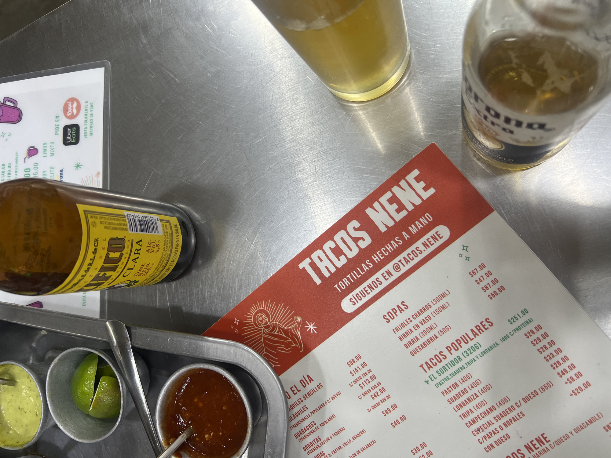 A tabletop scene at Tacos Nene, likely in Mexico City, features a menu displaying Mexican food items like "Milaquiles" and "Huaraches" and a Corona Extra beer bottle near a glass with beer. A tray with condiments, including a green sauce and limes, is also present. The menu advertises the restaurant's Instagram account "@tacos.nene" and lists food delivery services Uber Eats and Rappi.