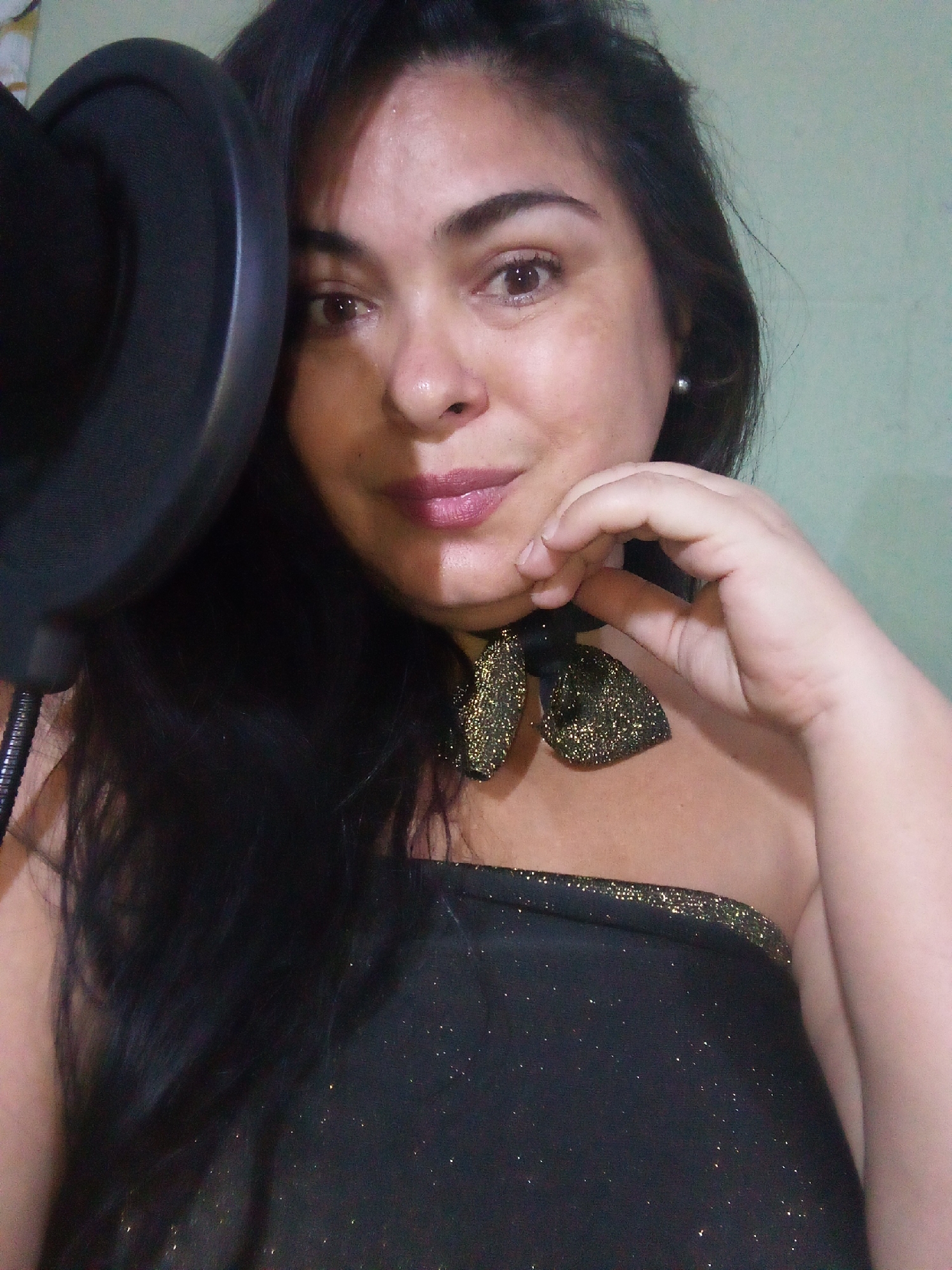 A woman poses indoors next to a microphone. She has dark hair, light skin, and is wearing a black strapless top with gold accents and a glittery bow tie. The background is a light greenish-blue. The city and country are not discernible from the image.