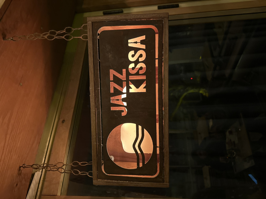 A rectangular, dark-colored sign with a light-brown wooden frame is suspended from a reddish-brown wooden ceiling by two sets of dark metal chains. The sign features illuminated cut-out letters spelling "JAZZ KISSA" in orange-amber light. To the left of the text, a circular illuminated design displays a stylized wave pattern and a partial reflection or image resembling a human profile. Below the sign, a large glass window reflects the interior of an establishment, showing dimly lit tables, chairs, and blurred figures. A small red light dot and a reflection of a green plant are also visible in the window. The establishment is identified as Jazz Kissa, located at Allenby Street 119, Tel Aviv-Yafo, Israel.