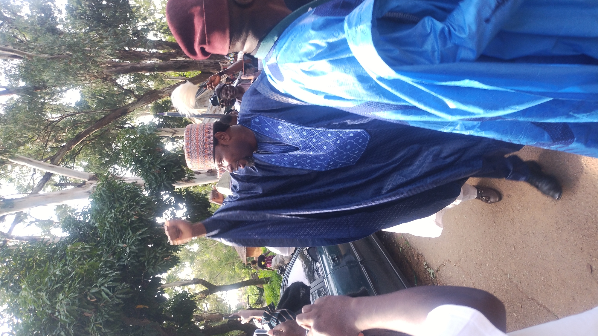 The media file shows a group of people gathered outdoors, with a focus on a man in the foreground wearing a dark blue, intricately patterned robe and a traditional cap. The man appears to be speaking or gesturing, with his fist raised in the air, suggesting an animated discussion or a moment of emphasis. He is wearing glasses and has a slight smile.
Behind him, other individuals are visible, some also dressed in traditional attire. A large, bright blue fabric, possibly a tent or banner, is draped prominently in the background, obscuring some of the scene. The setting appears to be a natural environment with trees, suggesting an outdoor event. The ground is a dirt or gravel surface.
The lighting suggests it is daytime, likely late morning or afternoon. There are no clear indications of the weather, though the lighting implies it is not raining. The presence of multiple people in traditional clothing and the outdoor setting suggest a communal gathering or event, possibly a cultural or religious ceremony, or a political rally given the animated posture of the main subject. The context provided places the scene in Jos, Nigeria. There is no visible text in the image.