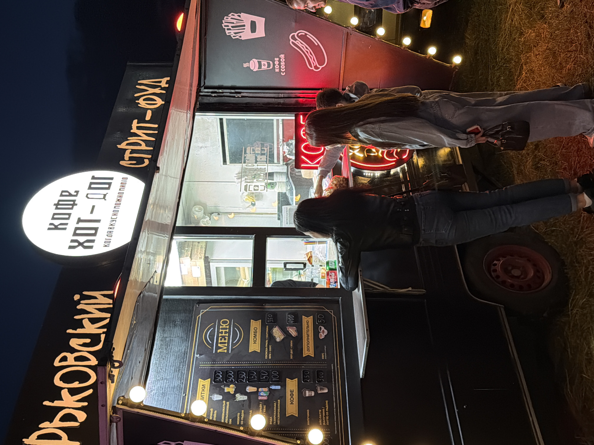 The image depicts a food truck at night. The food truck is black with illuminated signage. The main sign above the serving window reads "КОФЕ ХОТ-ДОГ" in large white letters within a circle, and below that, "ВКУСНО ПОЖИВНЕЮ". To the right of this sign, also illuminated, is "СТРИТ-ФУД". A menu is displayed on the left side of the serving window. It includes categories like "ВИТКИ", "МЕНЮ", "КОМБО", and "КОФЕ". Various items and prices are listed, such as " МОХИТО СО ЛЬДОМ" for 250, "КОМБО СОСИСКА-ГРИЛЬ + КАРТОШКА ФРИ + СОУС" for 350, and "КОФЕ КАПУЧИНО" for 250. Two women are standing at the serving window, appearing to order food. One woman is wearing a black leather jacket and jeans, while the other is in a denim jumpsuit. A third person, partially visible on the right, appears to be a man. The background is dark, indicating it is nighttime, and the ground appears to be covered in hay or dry grass. The food truck is brightly lit by internal lights and external bulbs along its side.