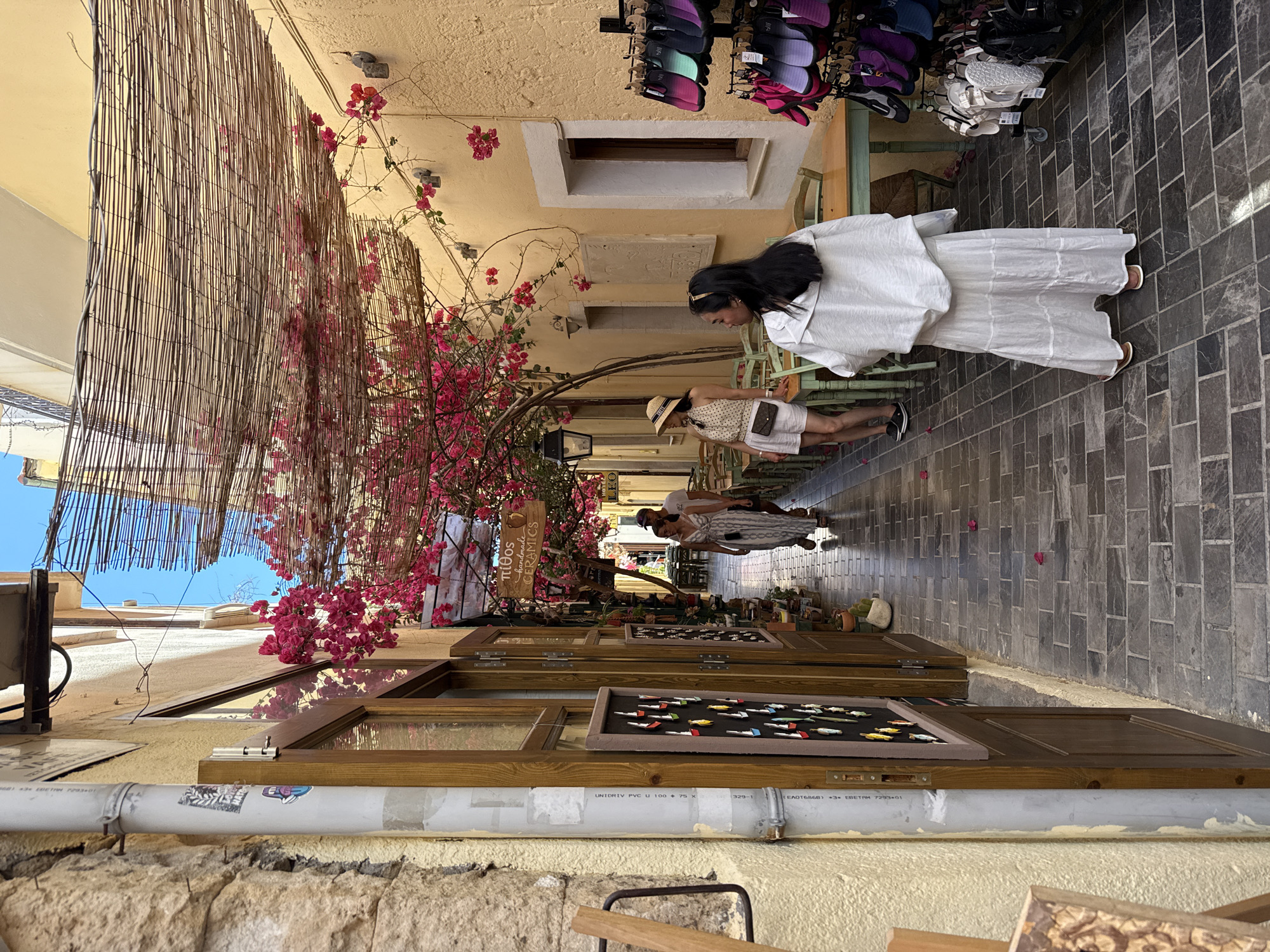 A narrow, cobblestone alleyway in the city of Rethymno, Greece, is lined with buildings adorned with blooming pink bougainvillea. A woven bamboo canopy shades the walkway. On the right, shelves display an array of colorful flip-flops and sandals. Tables and chairs are set up outside a building on the left, likely a cafe or restaurant. A woman wearing a white dress and headscarf stands facing away from the camera, while another woman in a straw hat and patterned top is visible further down the alley. The stone and yellow-painted walls of the buildings create a rustic atmosphere.
