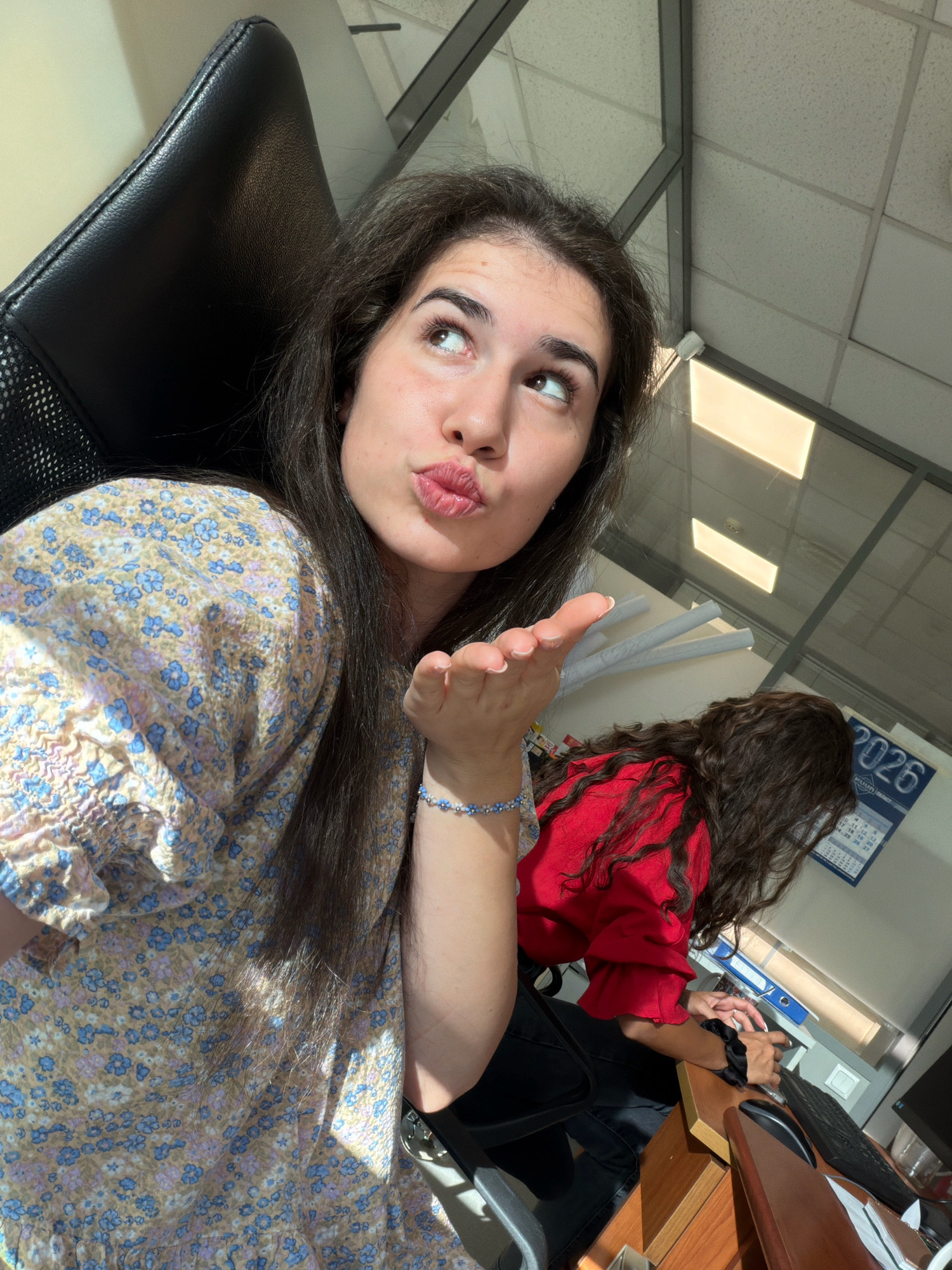 A young woman with dark, wavy hair is seated in an office chair, looking upwards and blowing a kiss towards the camera. She is wearing a light-colored dress with a floral pattern. Her right hand is extended, palm facing the camera, in a kissing gesture. Behind her, another woman with dark, curly hair, wearing a red top, is seated at a desk and working on a computer. A calendar with the year "2026" is visible on the wall behind the second woman. The environment appears to be a typical office setting with fluorescent lighting in the ceiling. The sunlight streaming in from a window casts shadows on the scene.