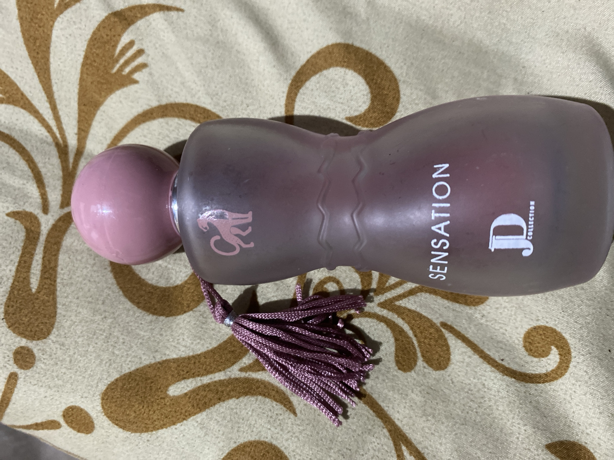 The image shows a frosted glass perfume bottle with a pink spherical cap and a pink tassel attached to its neck. The bottle is slightly curved and has the word "SENSATION" printed in white letters on its side, along with a stylized "JD COLLECTION" logo. A pink silhouette of a leaping animal, possibly a cheetah or a panther, is also visible on the upper part of the bottle. The perfume bottle is placed on a patterned fabric with a tan background and abstract brown motifs. The image was taken in Ta', Indonesia.