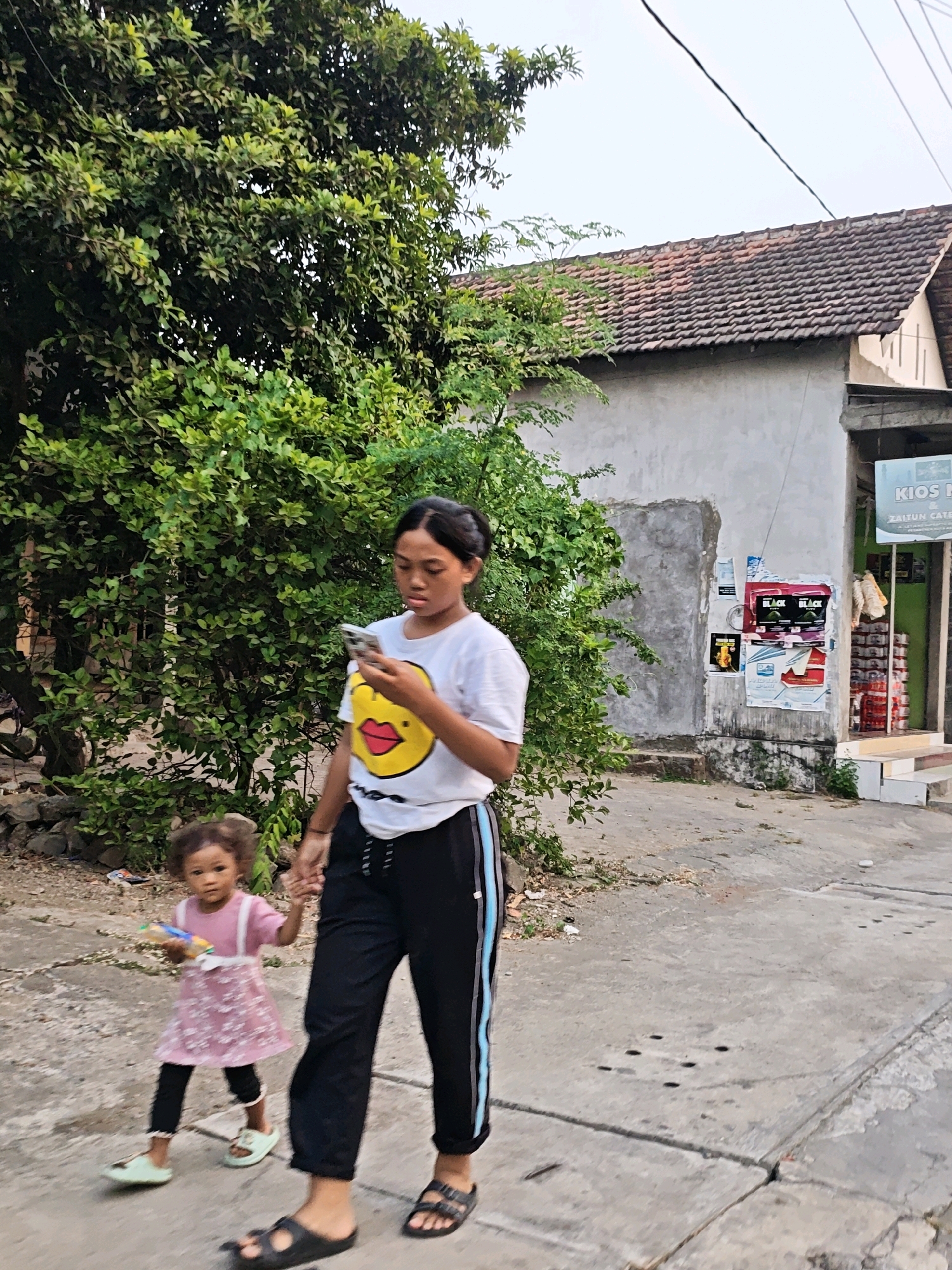 A teenage female, wearing a white t-shirt with a yellow duck graphic and black pants with a blue stripe, walks on a concrete sidewalk. She is holding a smartphone in her right hand and looking at it. A young girl, wearing a pink dress and black leggings, walks to her left, holding the teenager's hand. The girl is holding a toy in her left hand. They are walking in front of a building with a tiled roof and posters on the wall. Lush green trees are to the left of the building. The scene appears to be a residential street in Kediri City, Indonesia.