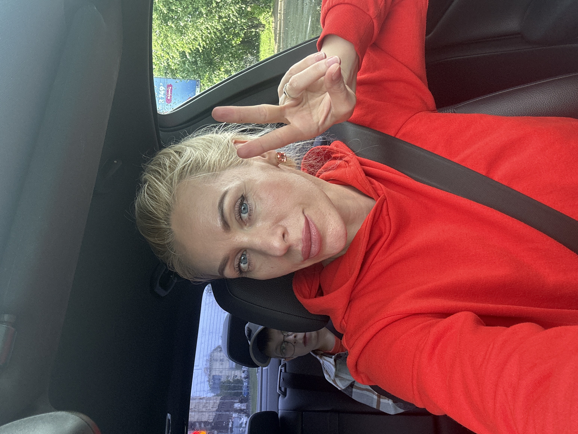 A blonde woman in a red hoodie takes a selfie from the driver's seat of a car. She is making a peace sign with her right hand, which has a ring on her ring finger. She is wearing an earring on her left ear. In the back seat, a young boy wearing glasses and a cap looks forward. The car is on a street in Saint Petersburg, Russia, with trees and buildings visible outside the windows.