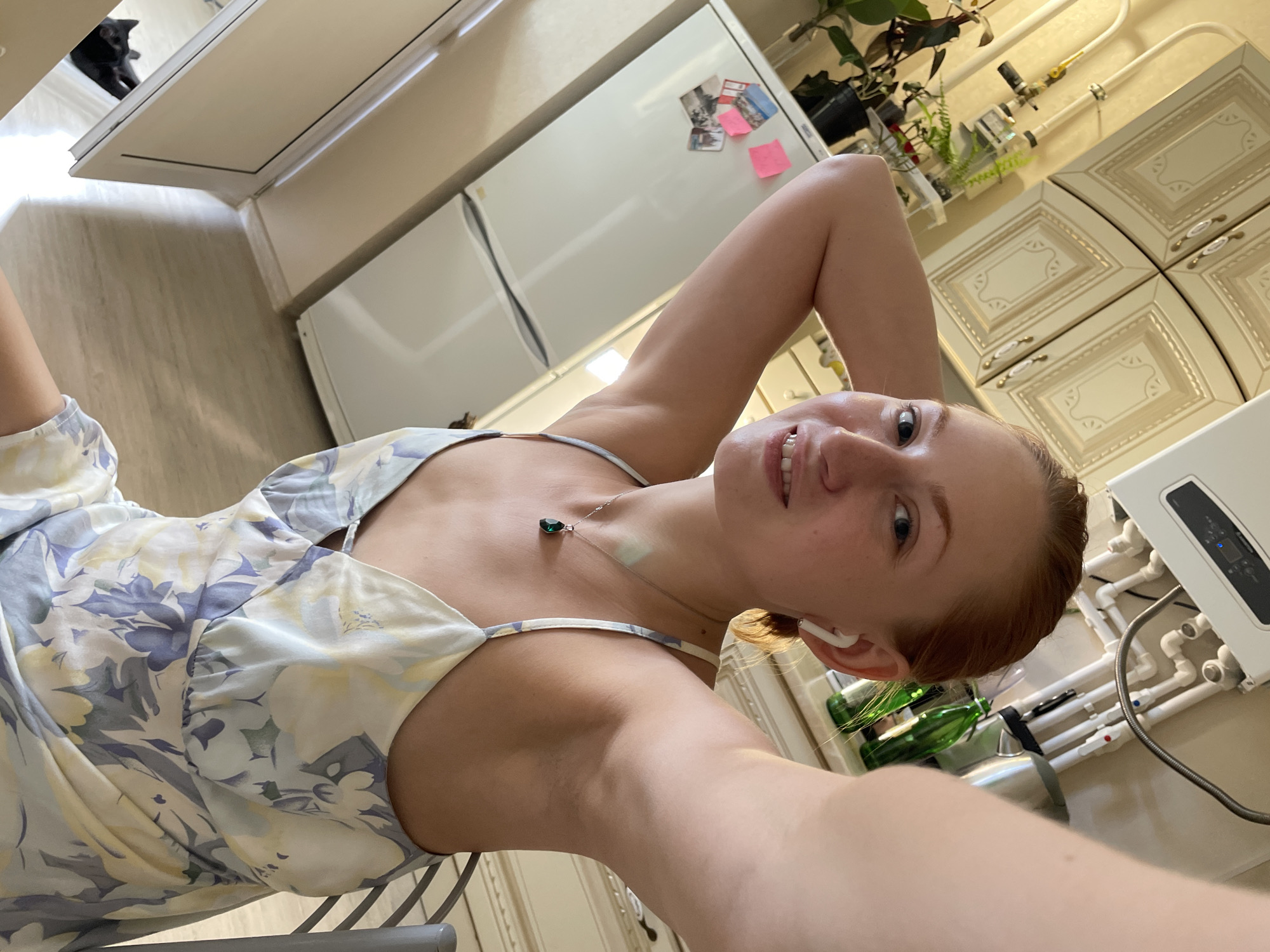 A young woman with red hair and fair skin is taking a selfie in what appears to be a kitchen. She is wearing a light-colored floral dress with thin straps and a green pendant necklace. She has white earbuds in her ears and is smiling at the camera, with her hands behind her head. To her left is a white refrigerator with some photos and sticky notes attached. Above the refrigerator, there are cream-colored kitchen cabinets and some plumbing. To her right, a dark-colored cat is visible near the floor. The flooring appears to be light wood. There is no indication of the city of Anapa, Russia, in the image itself.