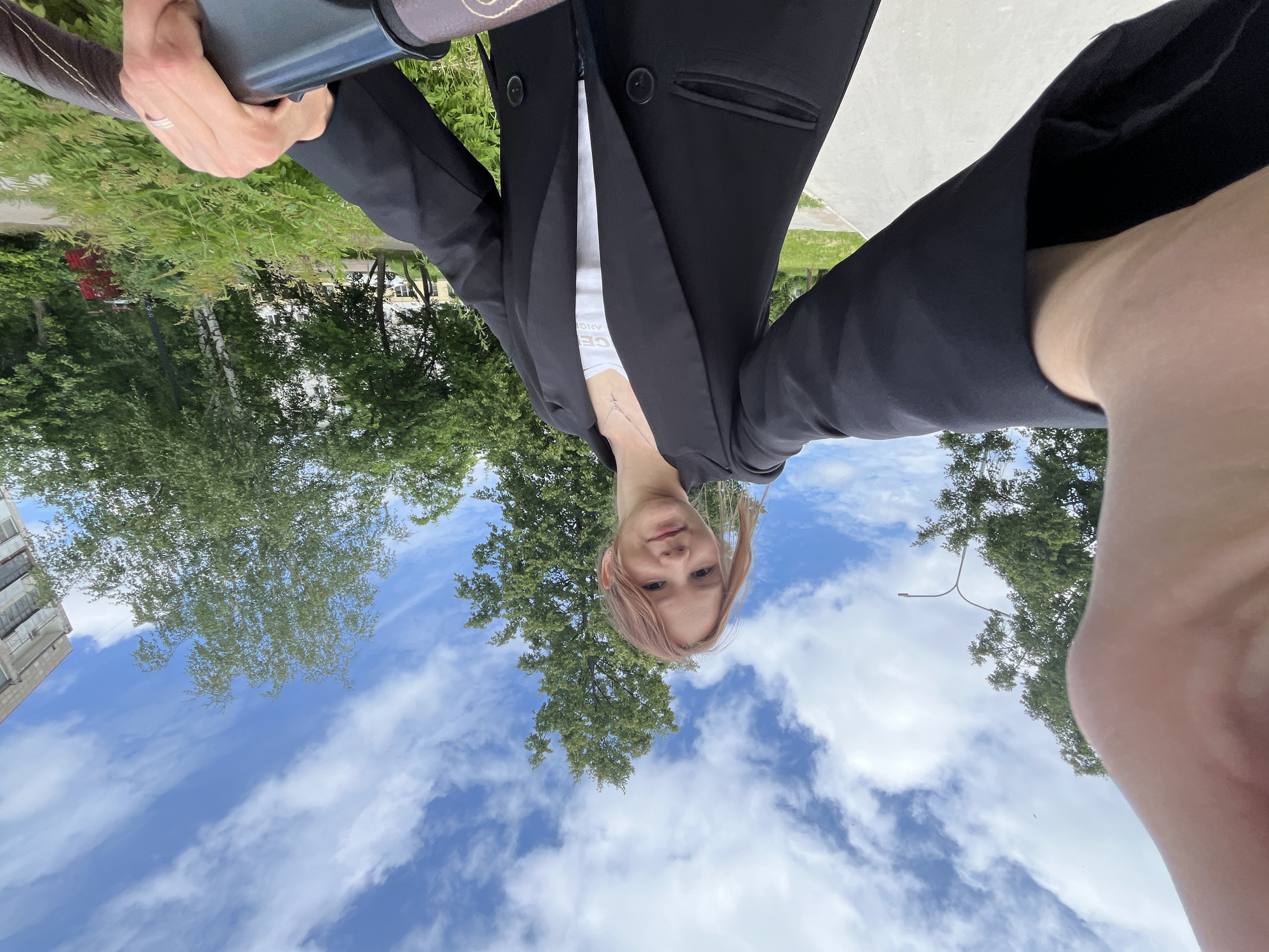 A young woman with reddish hair, wearing a black blazer over a white t-shirt and a silver cross necklace, is taking a selfie outdoors. She is holding the camera with her left hand, which is positioned in the foreground, obscuring part of the frame. The woman is smiling and looking at the camera. Behind her, a tree with green foliage stands tall against a blue sky with scattered white clouds. To the right, a building with balconies is visible, and in the lower right corner, a person's hand with a ring is gripping a brown handlebar, possibly of a stroller or bicycle. The scene appears to be a park or residential area in Chaikovsky, Russia, with a paved path in the foreground and lush greenery surrounding the area.