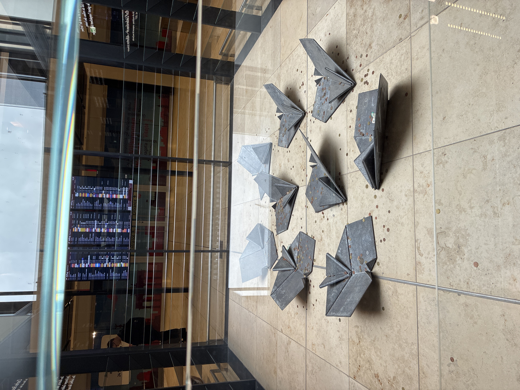 The image depicts an indoor scene at Berlin Brandenburg Airport (BER) in Schönefeld, Germany. The foreground features several metal origami-style sculptures placed on a tiled floor. Scattered around these sculptures are numerous coins, suggesting they might be part of a wishing well or donation setup. In the background, a departure information board displays flight details. A person wearing glasses and a backpack stands near the information board, looking at it. The overall environment appears to be a public space within an airport terminal, characterized by polished floors, modern architecture, and displays. A reflective glass barrier separates the viewer from the scene.