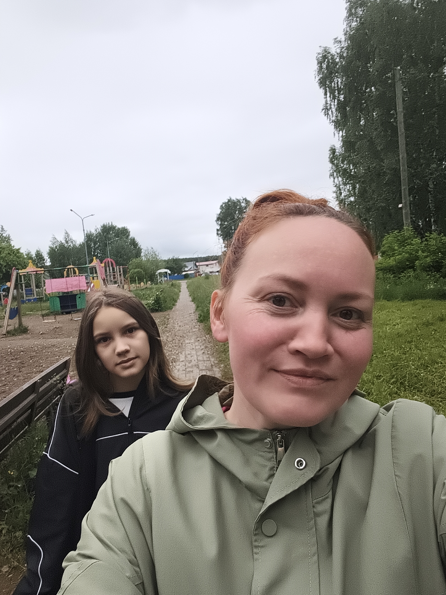 The image depicts an outdoor selfie featuring two females in what appears to be a rural or suburban setting under an overcast sky.
In the foreground, an adult female is positioned on the right, taking the photo. She has reddish-brown hair pulled back, a light complexion, and is smiling faintly with visible lines around her eyes. She wears an olive green hooded jacket with a prominent silver snap button on the collar.
Behind and to the left of the foreground subject, a younger female (appearing to be a teenager or pre-teen) stands, looking directly at the camera with a neutral expression. She has long, straight, dark brown hair and is wearing a black zip-up jacket with white piping details over a white garment.
The background reveals a winding, unpaved or brick-paved path leading into the distance. To the left of the path, a colorful children's playground with swings, slides, and various structures (pink, yellow, blue, green elements) is visible on a dirt ground. A streetlight with a curved arm stands nearby. Beyond the path and playground, lush green trees dominate the landscape, with some taller deciduous trees on the right. In the far distance, faint residential structures, including one with a red roof, can be seen amidst dense foliage. The ground in the foreground and along the right side of the path is covered in green grass and weeds.
The location cannot be identified as a specific city but appears to be a community area or park in a natural, less urbanized environment.