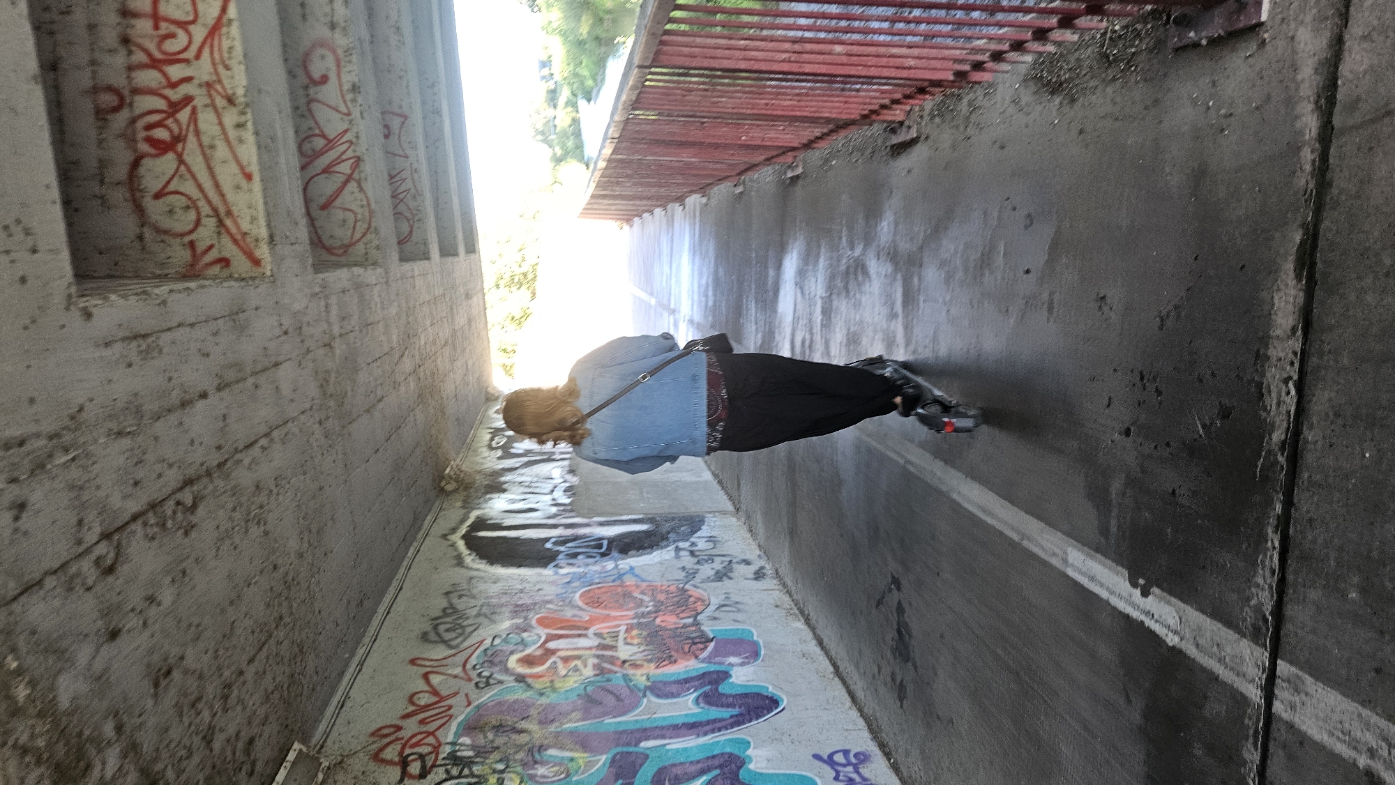 A person rides an electric scooter along a concrete path under a bridge adorned with graffiti. The path is wet, and a white line marks its edge. To the right, a red metal railing separates the path from a body of water and greenery. The bridge's concrete ceiling also displays graffiti. The person, viewed from behind, wears a denim jacket and black pants, and carries a black bag. The image appears to be taken in Prešov, Slovakia.
