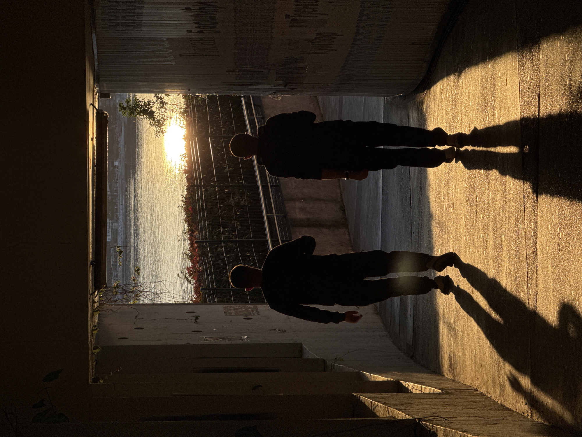 The image shows a figure and its shadow in an urban tunnel or underpass in what is likely Boreti, Montenegro, based on the environment. The image is taken from a low perspective, near the ground.
The figure, silhouetted against a bright, sunlit view of water (possibly the Adriatic Sea), appears to be a man with a shaved head, wearing dark clothing consisting of a long-sleeved top and trousers. The man's shadow is cast on the right side of the image onto the tunnel wall and the ground.
In the background, the tunnel exit frames a body of water with sunlight reflecting on it. There is vegetation visible above the water line, suggesting a shoreline. The tunnel itself is constructed of concrete. The walls of the tunnel show signs of wear and contain graffiti or markings. The ground appears to be paved. The light source from the tunnel exit creates a strong contrast between the dark tunnel interior and the bright exterior. The image is rotated counterclockwise by 90 degrees.