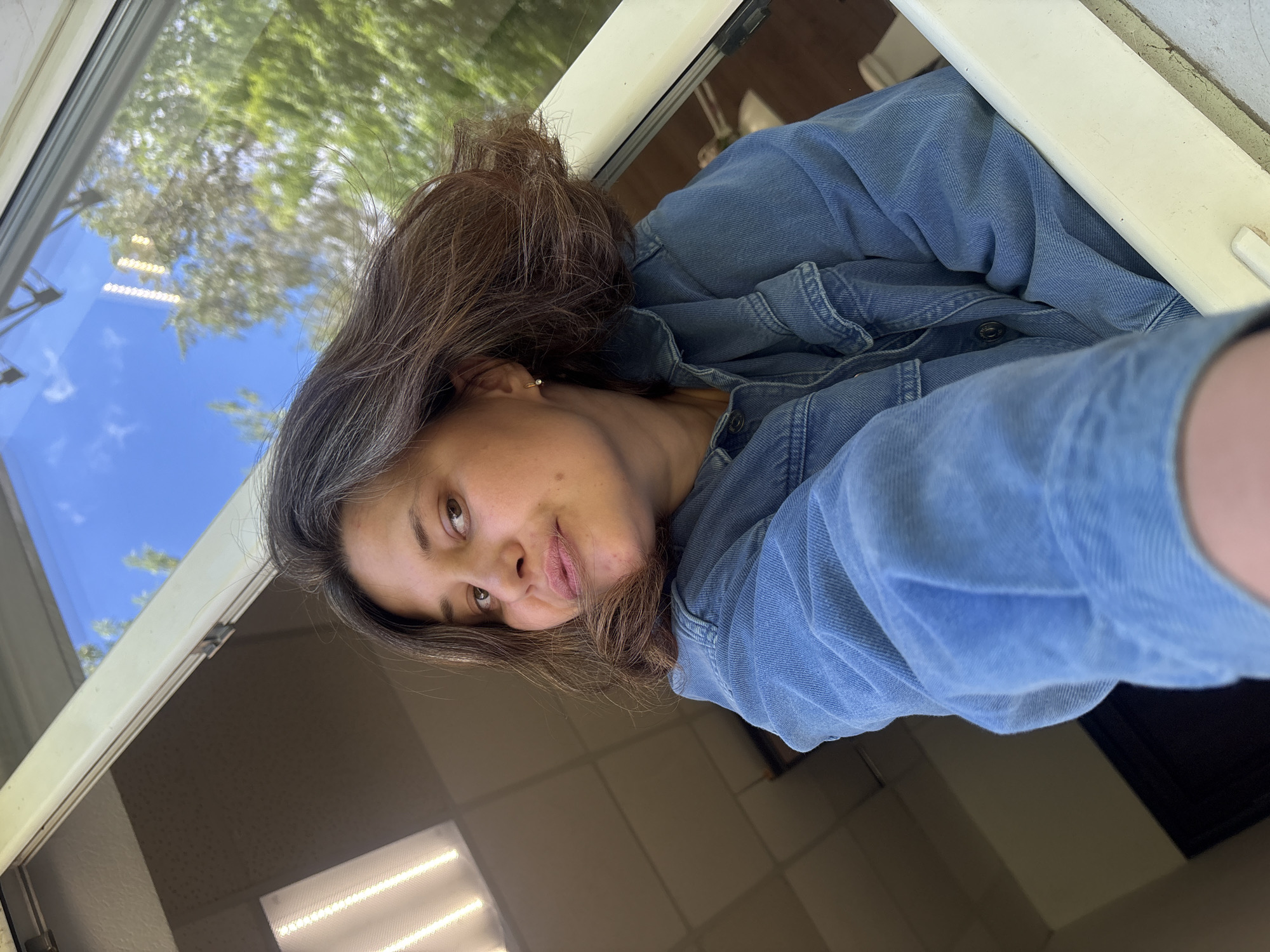 This is a close-up, rotated selfie of a young woman wearing a denim jacket. She has brown hair with some gray streaks, and her head is tilted to her right, with her gaze directed towards the camera. She is wearing a small hoop earring in her visible ear. The background shows a window with a reflection of blue sky, green trees, and some overhead lights. The interior of the room behind her is visible, with a tiled ceiling and fluorescent lighting. The woman's expression appears neutral or slightly pensive. The image seems to have been taken during daylight, indicated by the bright sky and outdoor reflections. The overall impression is that of a casual, personal snapshot taken indoors near a window.