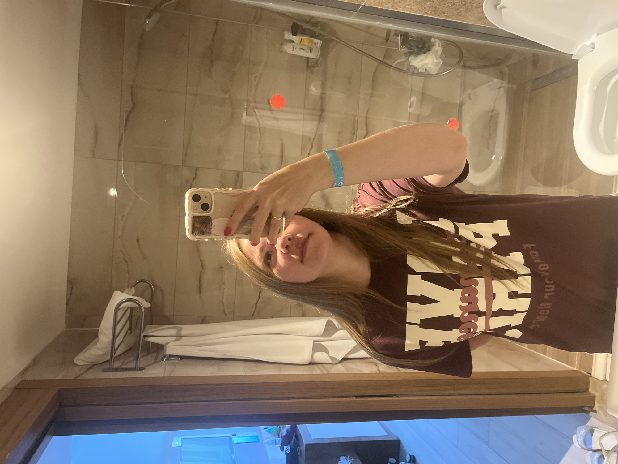 A young woman with long blonde hair is taking a mirror selfie in a modern hotel bathroom. She is wearing a maroon t-shirt with large white lettering and a blue wristband. Her right hand is holding a beige phone with a floral case, and her left arm is bent at the elbow. The bathroom features marble-patterned tiles on the walls, a glass shower enclosure with a shower head, a towel rack with white towels, and a white toilet. To the left of the frame, a wooden door opens to reveal a brightly lit room with blue accents and a white counter. The overall ambiance suggests a hotel in Alanya, Turkey, likely a resort or a vacation rental.