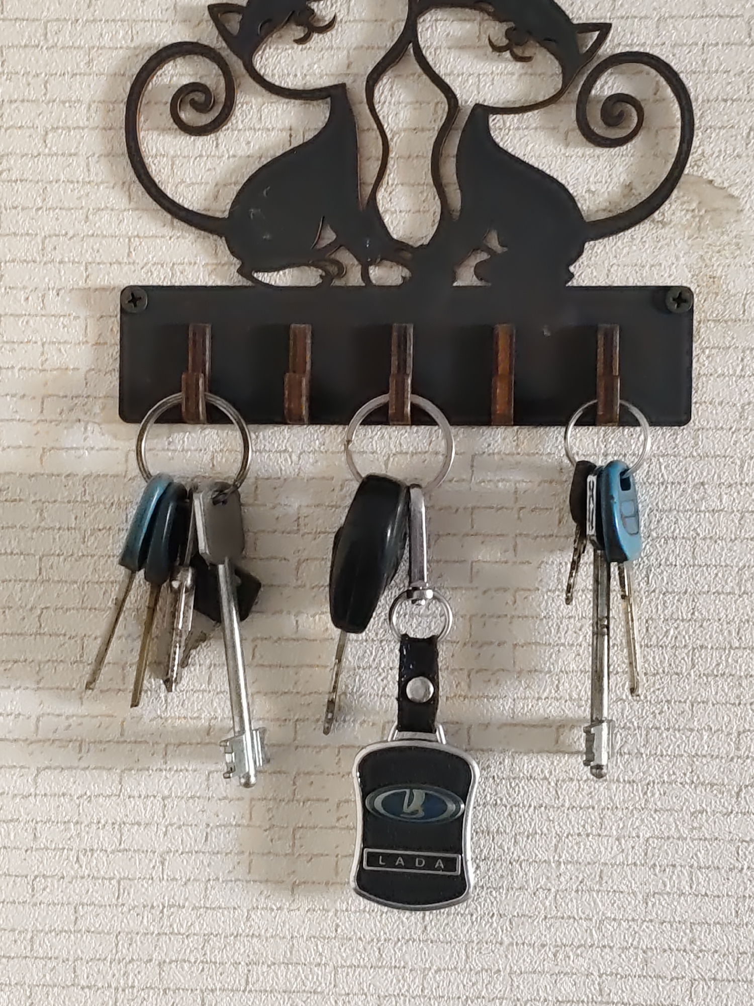A black metal key rack with a decorative design of two cats is mounted on a white brick-textured wall. The key rack has five hooks, and three sets of keys are hanging from the left three hooks. The leftmost set of keys has two keys with blue plastic heads and one silver key. The middle set of keys has one black plastic key fob with a silver emblem and text that reads "LADA" and one silver key. The rightmost set of keys has one blue plastic key and two silver keys. The image appears to be taken indoors. There is no visible indication of the location being Nizhnedonskoy, Russia.