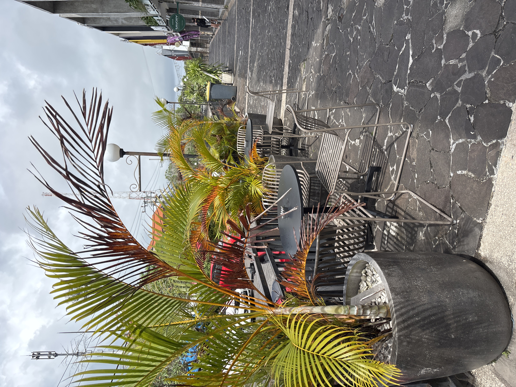 The image depicts an outdoor cafe setting in Kuta Selatan, Indonesia. The foreground is dominated by a large, dark-colored planter containing a palm tree with a mix of green and brown fronds. The planter is filled with white pebbles. Behind the planter, there are several small, round tables and metal chairs arranged along a stone-paved walkway. Further back, there's a trash can and glimpses of other plants and objects. In the background, there are buildings and overhead wires, and a cloudy sky above. The scene appears to be taken during the daytime.