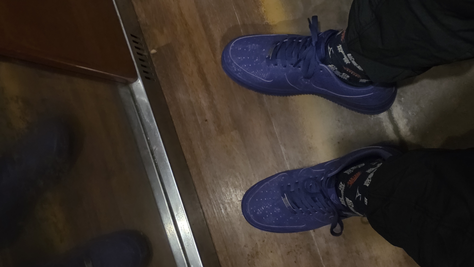 A person wearing matching dark blue sneakers and patterned socks is standing on a wooden floor. The person is also wearing dark-colored pants. A reflective surface, possibly part of an elevator, is visible on the left side of the image. The setting appears to be indoors, with a wooden floor. There is no information present in the picture to indicate this location is in Varna, Bulgaria.