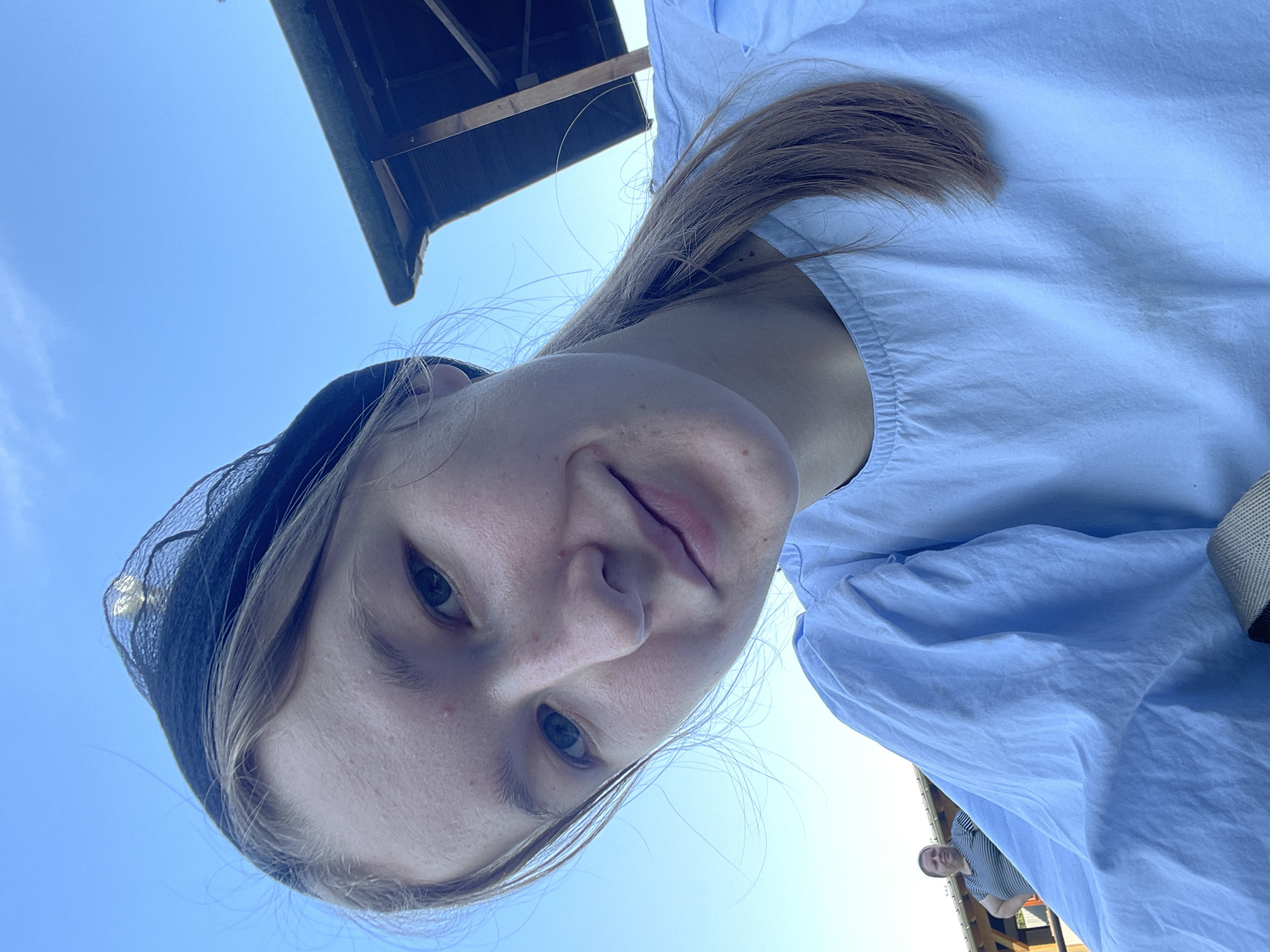 A young woman with light brown hair and a black head covering is taking a selfie outdoors. She is wearing a light blue ruffled shirt. In the background, there is a wooden structure, a clear blue sky, and another woman standing further away. The lighting suggests it is daytime. The exact location in Beltershain, Germany is not discernible from the image.
