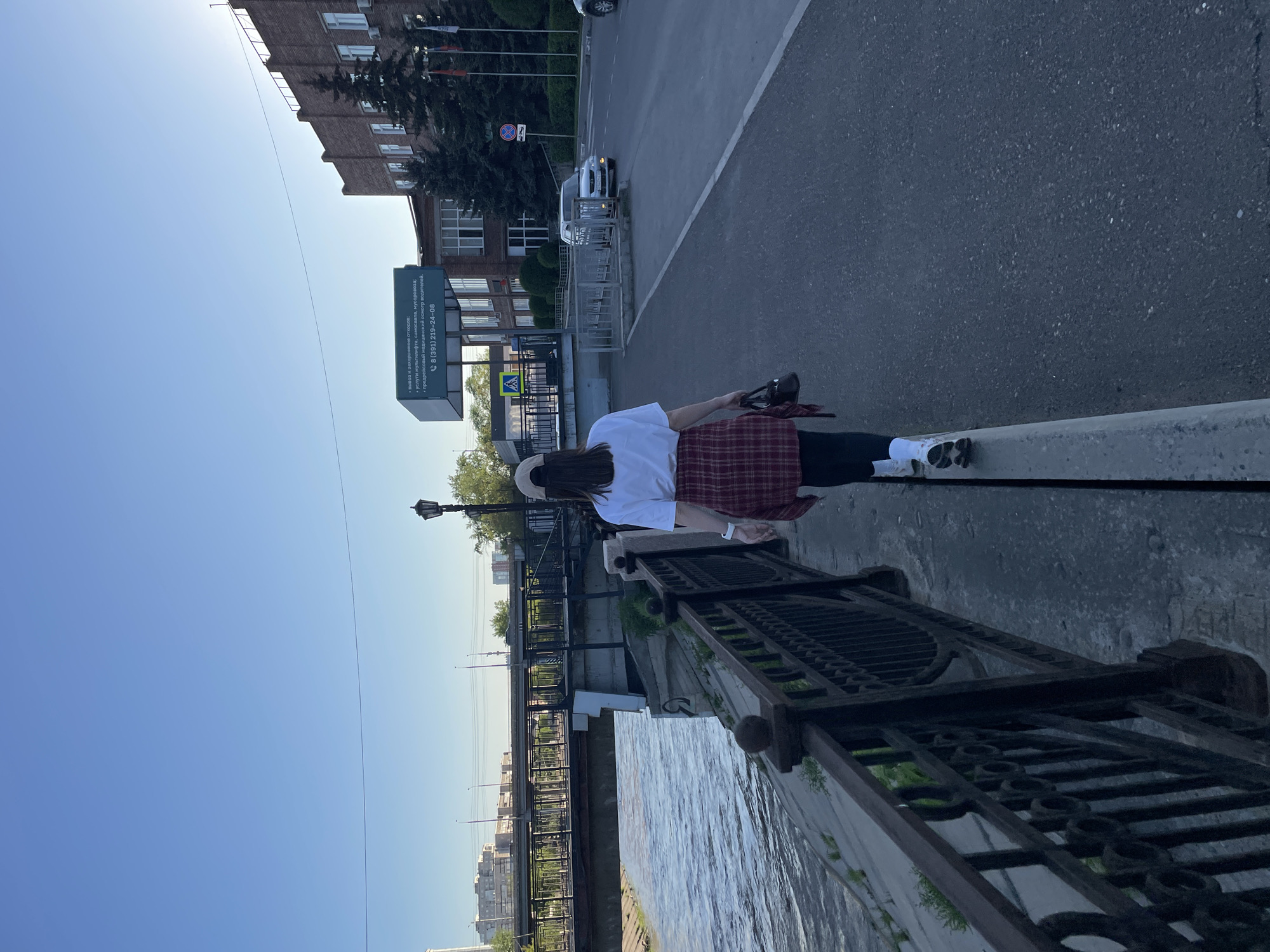 The image depicts a woman standing on a bridge with an ornate metal railing in Krasnoyarsk, Russia. The view from the bridge includes a body of water (likely the Yenisei River), with a city skyline visible in the distance. A pedestrian walkway runs parallel to the river. The street has a building that has a sign: " вывоз и захоронение отходов; *услуги мультилифта, самосвала, мусоровоза; *предрейсовый медицинский осмотр водителей. 8 (391) 219-24-08." The image is taken during the day.