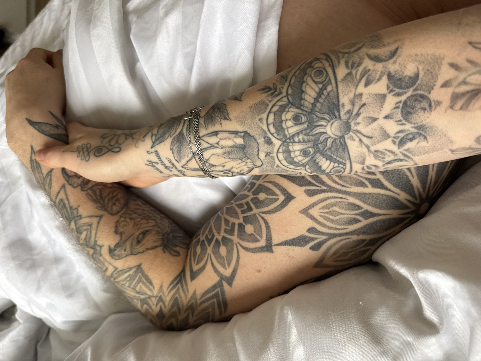 The image displays a close-up, overhead view of two individuals' forearms and hands resting on white bed sheets with subtle striped patterns.
The arm on the left, exhibiting some hair, is extensively covered in black and grey tattoos. Visible designs include a prominent wolf-like animal head with open jaws, various geometric patterns, and complex mandala-style motifs.
The arm on the right, appearing less hairy and more slender, is also extensively tattooed in black and grey. Noteworthy designs include a large butterfly or moth with a central sun emblem, crystal formations, a series of crescent moon phases, floral elements, a stylized unalome-like symbol, and partially visible Cyrillic script, which appears to read "за руку лучшей..." (by the hand of the best...). A silver-colored chain bracelet with an initial "M" charm is worn on this forearm.
The two arms are intertwined, with the right hand gently clasped over the lower forearm/wrist area of the left arm. This suggests a close physical interaction. The immediate environment is an indoor setting, specifically a bed. There are no identifiable visual elements within the image to specify a location in the city of Rostov-on-Don, Russia.