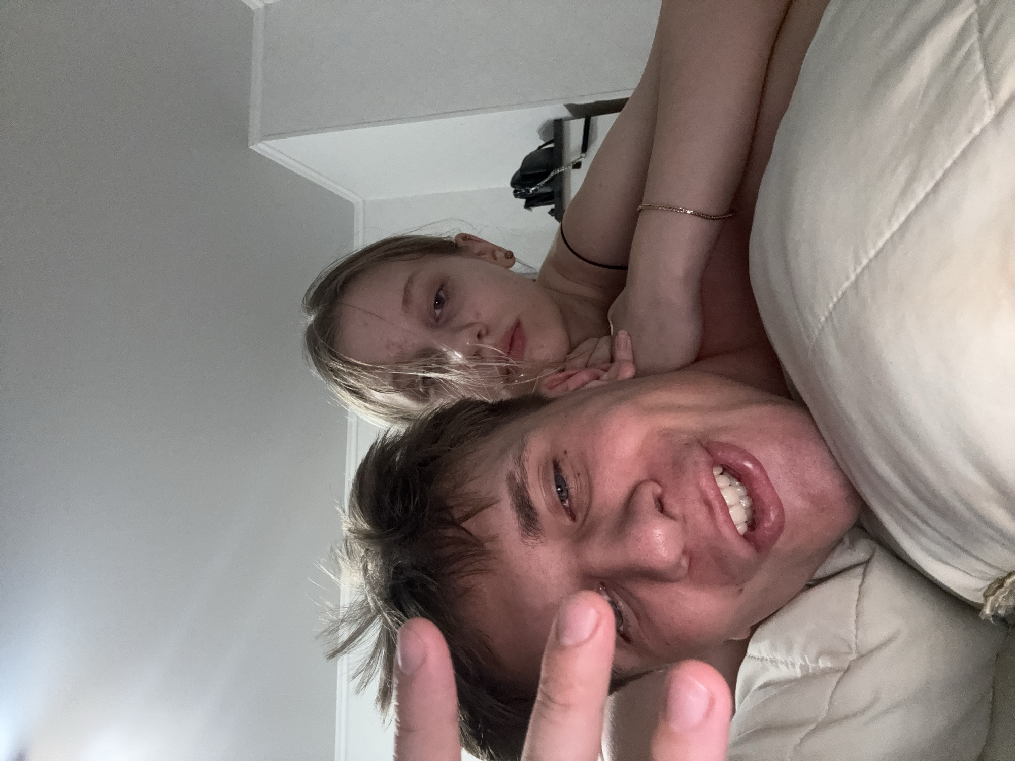The image is a selfie taken by a man lying in bed. A young woman is behind him, also in bed. The man is smiling and making a "peace sign" with his fingers. The woman is looking at the camera with a neutral expression. The room appears to be a bedroom, with a light-colored wall and ceiling visible. There is a pillow visible under the man's head, and a duvet is also present. A black bag and a shelf are visible in the background behind the woman. The lighting suggests it may be nighttime or a dimly lit room. The image does not provide enough information to determine a specific location within Tiraspol, Moldova.