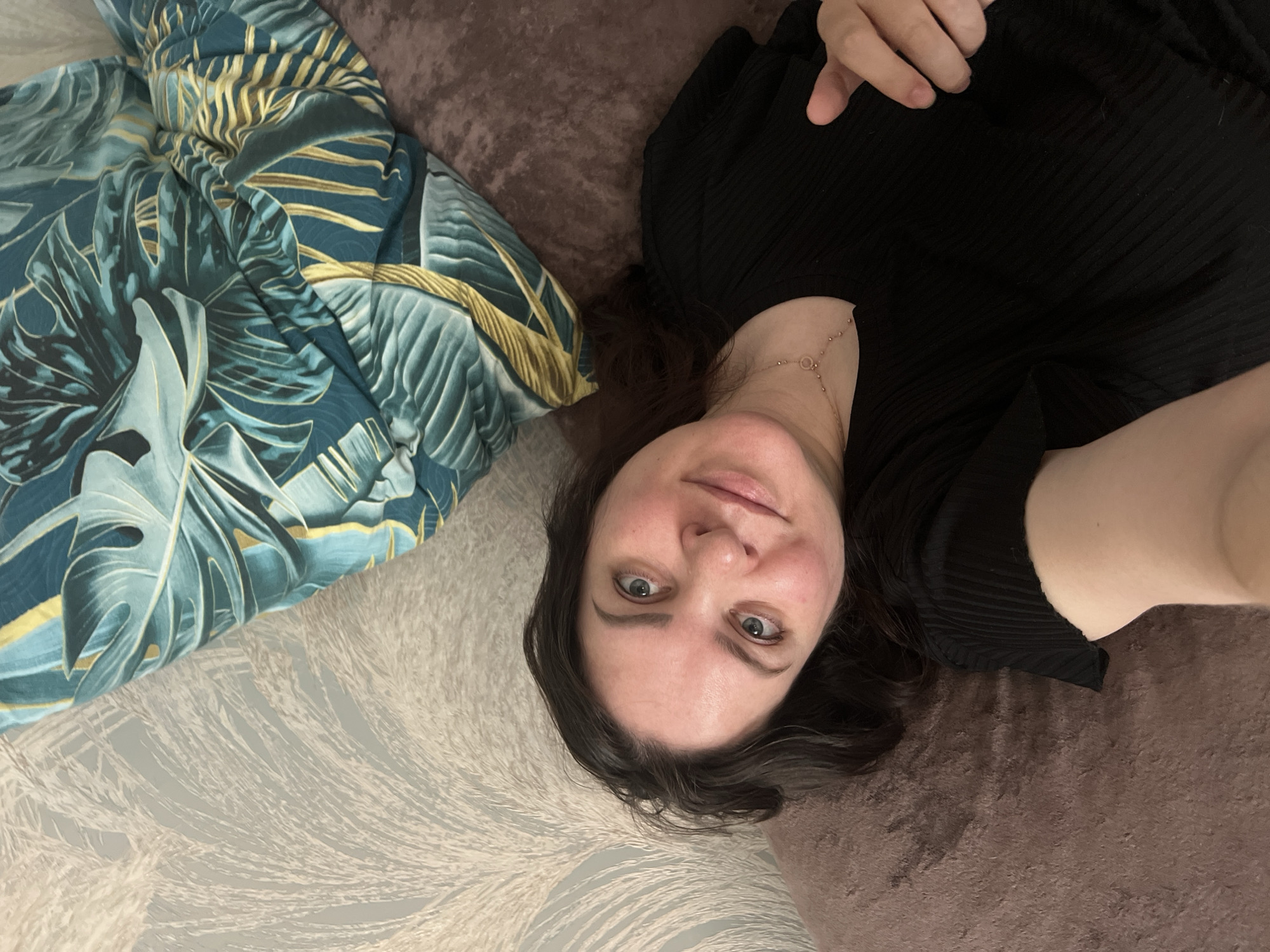 A woman is lying on a bed. She is wearing a black ribbed top and a delicate gold necklace. Her hair is dark and curly. She is looking directly at the camera with a slight smile. To her left is a patterned pillow with blue and gold monstera leaves. The bed has a textured, light gray and white patterned sheet on one side and a solid brown headboard or cushion on the other. The photograph appears to have been taken from above. The city and specific location of the image is not identifiable from this image.