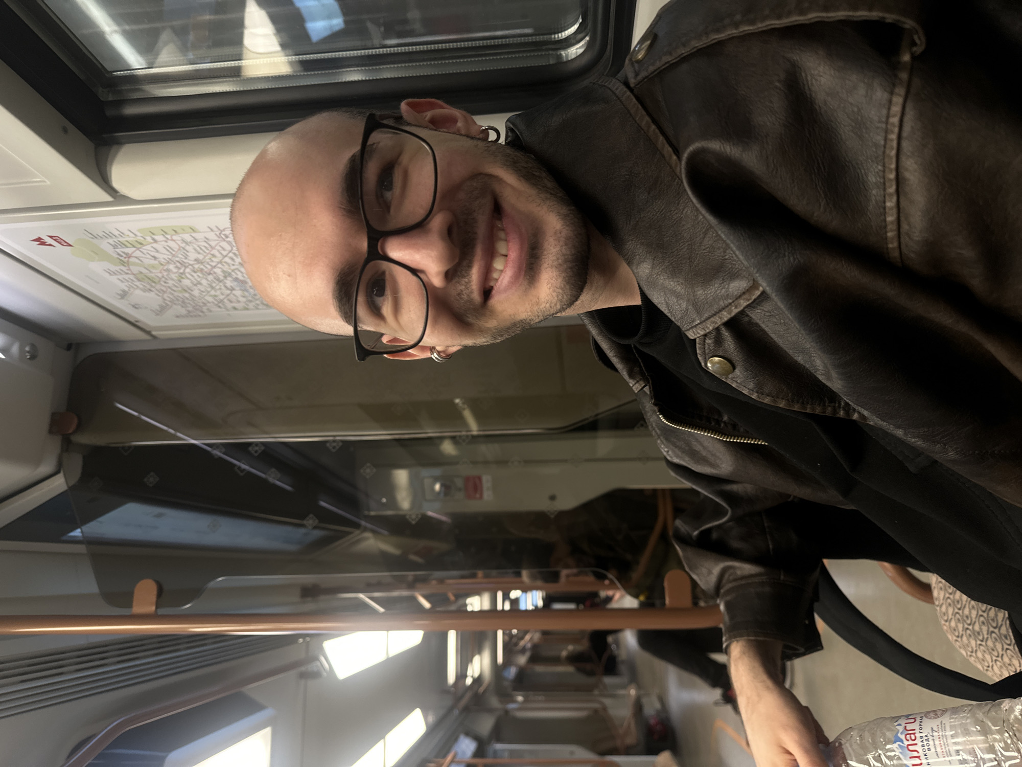 A smiling, bald man with glasses and two earrings is on a train. He wears a dark leather jacket over a black shirt. The train is a modern type, with a map displayed above the window. The man is holding a clear plastic water bottle. The train is in Moscow, Russia.