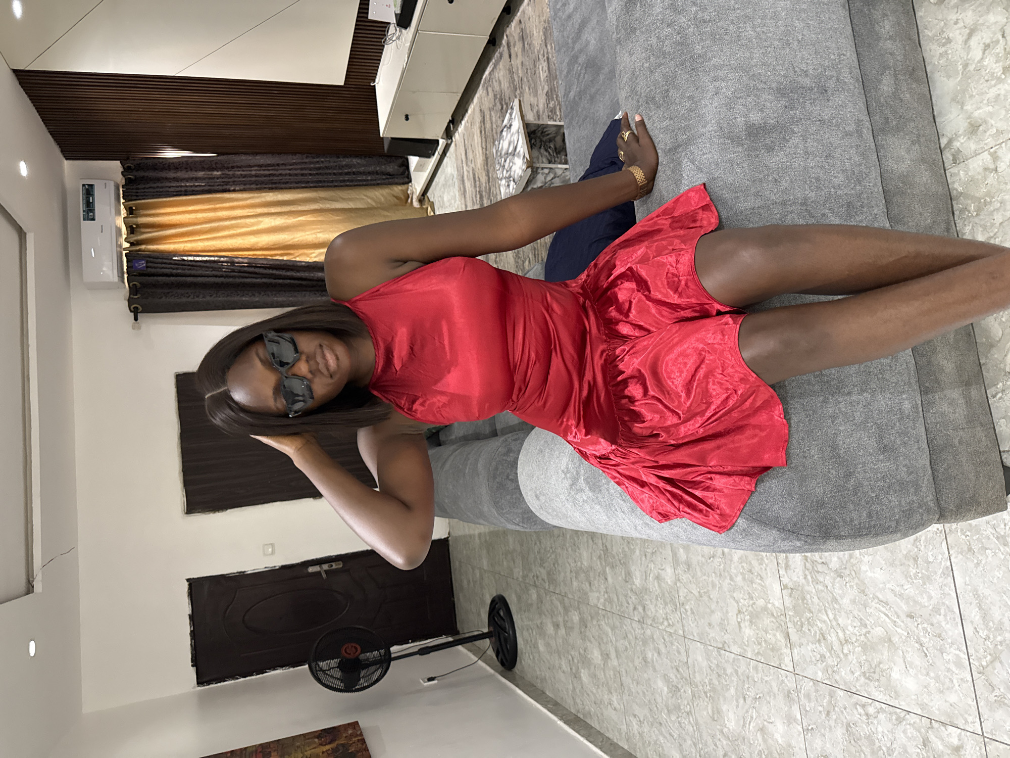 A woman wearing a red dress and sunglasses is reclining on a grey sofa indoors. The room features a dark wooden door, a black fan, and decorative curtains. In the background, there is a white cabinet and a glimpse of a patterned floor. The location is presumably a residential interior in Iddo Tudunwada, Nigeria.
