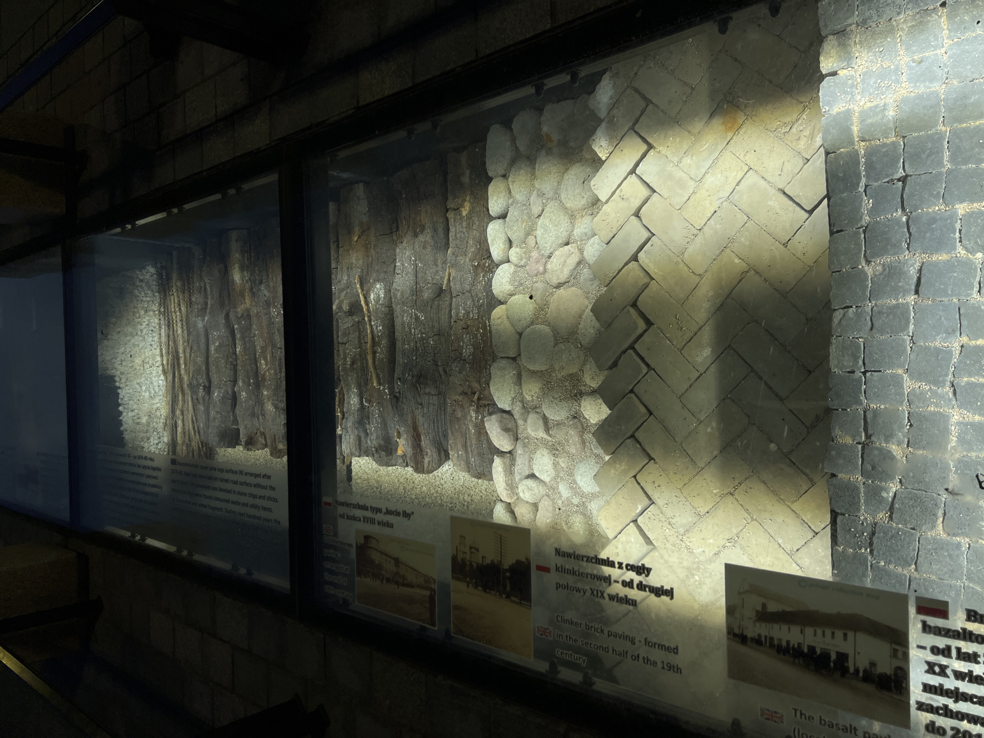 The image shows an archaeological exhibition displayed under glass flooring in Lublin, Poland. The exhibition features layers of historical pavement, including wooden logs, cobblestones, and brickwork. Information panels with text and historical photographs are visible alongside the display, providing context about the archaeological findings. The flooring is composed of different types of stone and brick, arranged in various patterns. The scene is illuminated, highlighting the preserved historical materials.