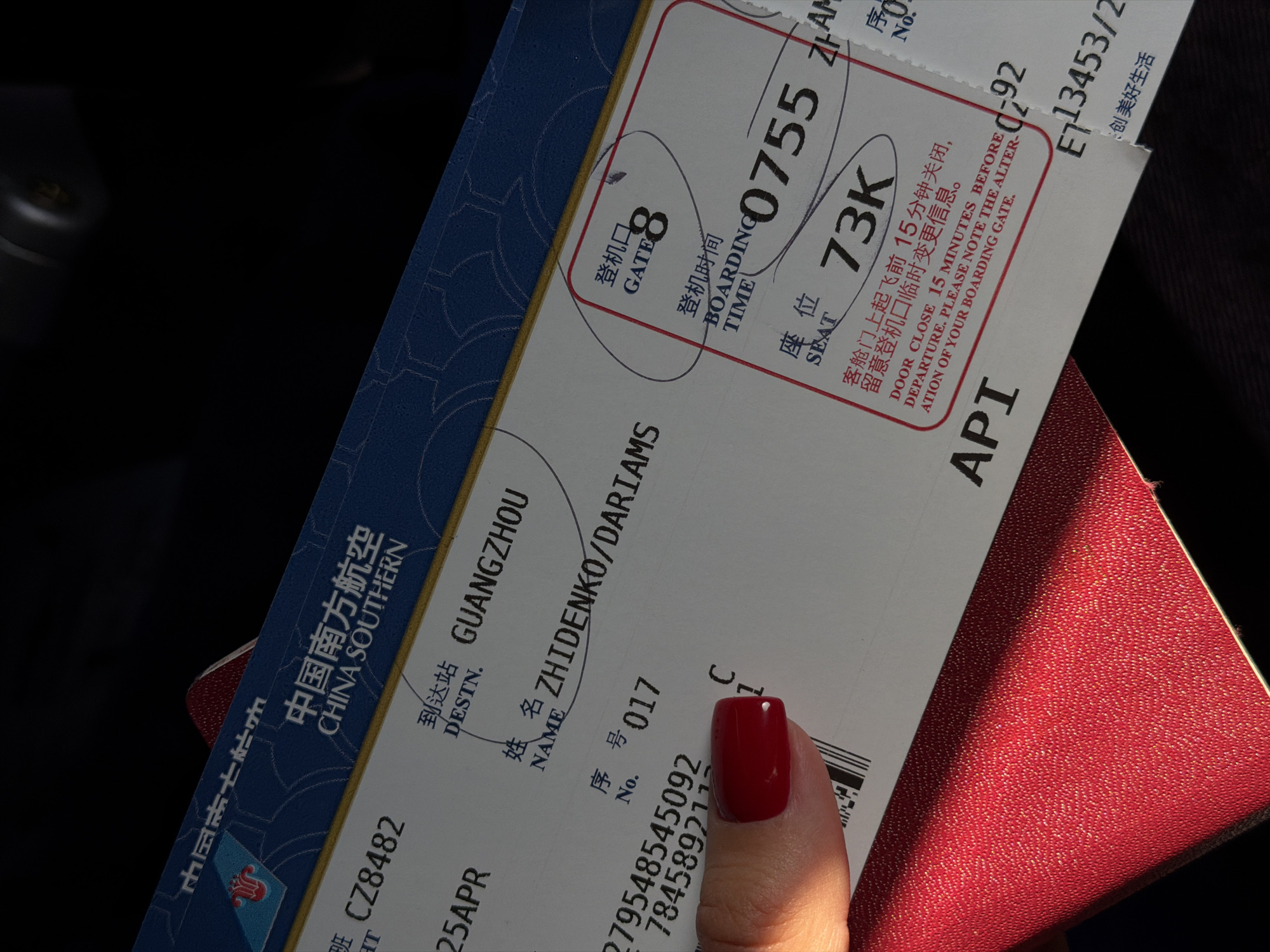 The image shows a boarding pass for a China Southern flight CZ8482 to Guangzhou (CAN) departing on April 25th. The passenger's name is ZHIDENKO/DARIAMS. The boarding gate is 8, and the boarding time is 07:55 AM. The seat number is 73K. The boarding pass is placed on top of a red passport. A hand with a red nail is partially visible, holding the boarding pass. The surrounding environment is dark. No location in Kuta, Indonesia, is visible.