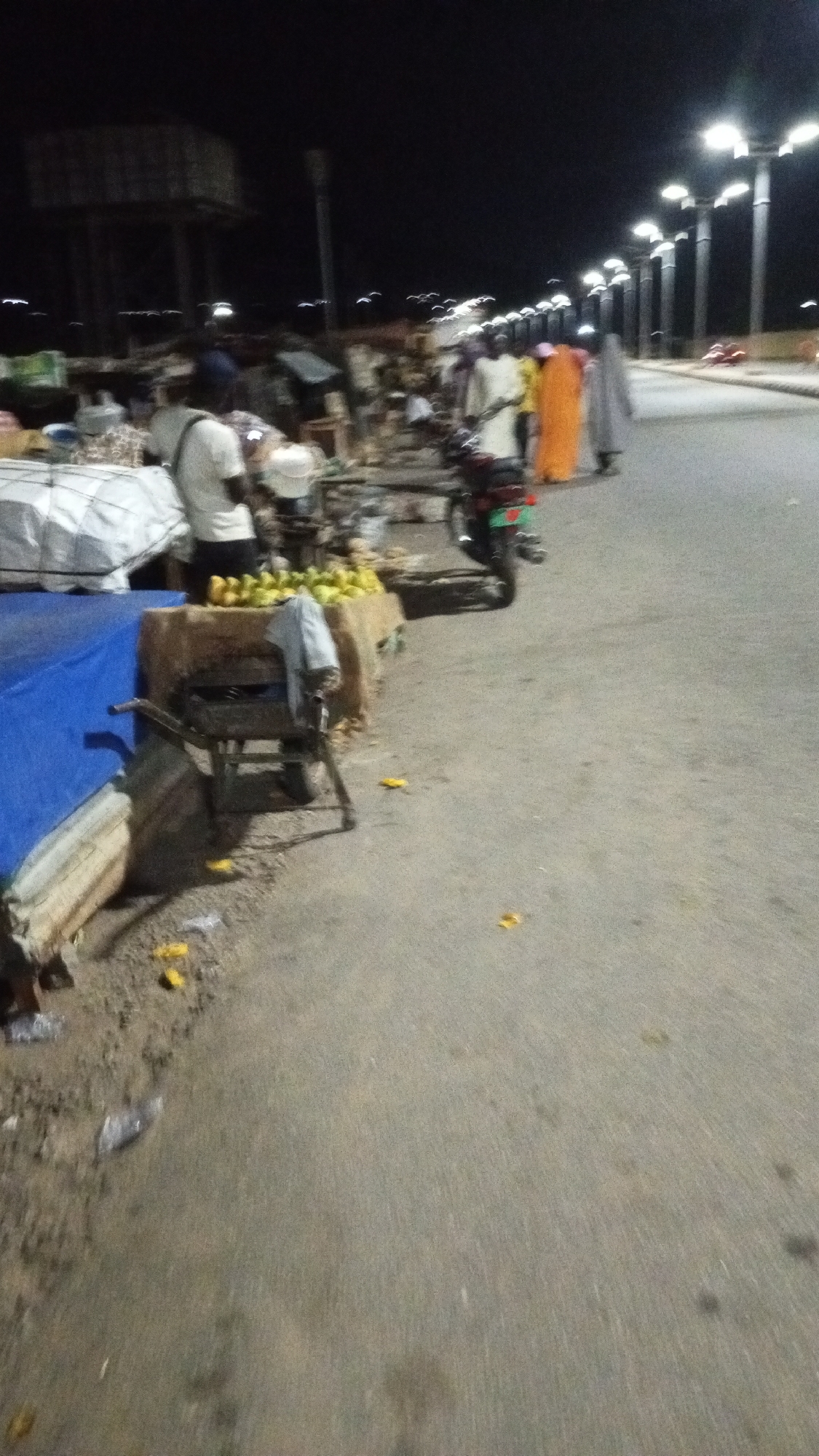 The image depicts an outdoor scene at night in Adankolo, Nigeria. Along the roadside, there is a market area with various stalls and vendors. Some individuals are walking along the road, with one wearing an orange robe. A motorcycle is parked near a stall selling mangoes, with a man standing nearby. There are street lights illuminating the area and a water tower is visible in the background. Trash and discarded mango peels litter the ground.