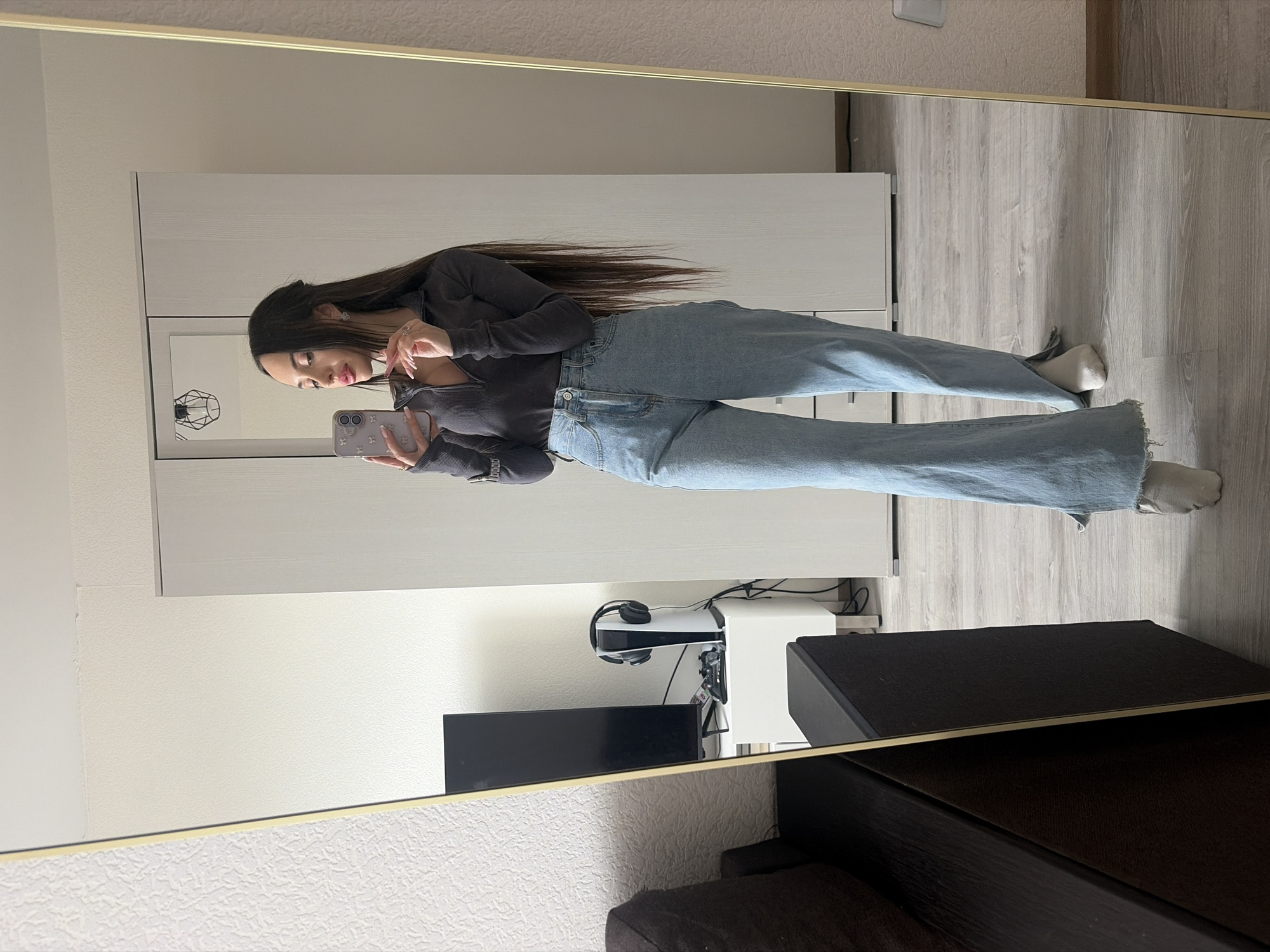 A woman stands in front of a mirror, holding a smartphone, presumably taking a selfie. She is wearing a long-sleeved, dark-colored top and light blue jeans. Her long brown hair is parted to the side. The reflection reveals a room with light-colored walls and flooring. Visible in the background are a closet or wardrobe, a TV, a Playstation 5, headphones, and a piece of dark furniture, possibly a dresser or side table. The scene takes place indoors. There is no evidence suggesting the location in the city of Saint Petersburg, Russia.