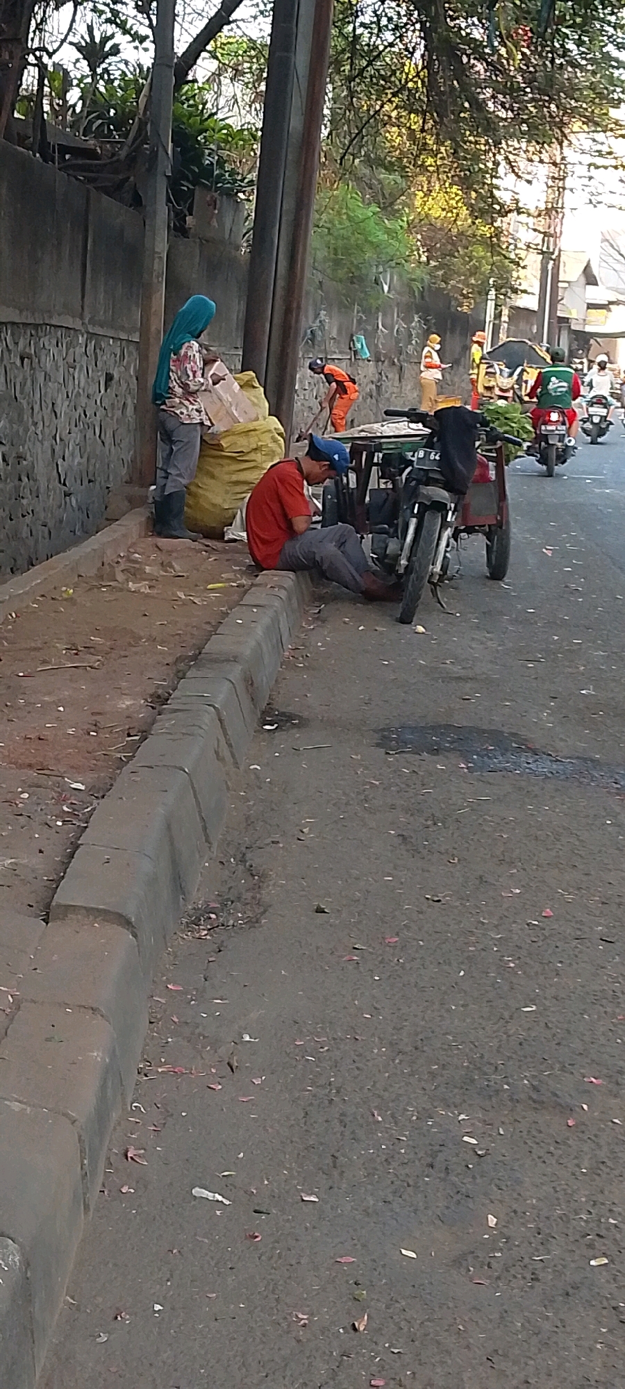 The image depicts a street scene in Bekasi, Indonesia, with a focus on individuals and their activities along the roadside. In the foreground, a man wearing a red shirt and a blue cap is seated on a concrete curb, appearing to be using a mobile phone. Beside him is a three-wheeled vehicle, likely a modified motorcycle with a cargo carrier, which is parked. Further back, two individuals are interacting near a large yellow bag and some debris on the side of the road. One of them appears to be wearing a blue headscarf and grey trousers, while the other is dressed in orange work attire, possibly engaged in cleaning or waste collection. In the background, several other individuals in similar orange work clothes are visible, some holding shovels, suggesting a work crew. The environment shows a paved road with scattered debris and fallen leaves, alongside a stone wall and vegetation. A utility pole is also prominently featured in the scene. The overall impression is one of urban street life, with elements of manual labor and daily activities taking place.