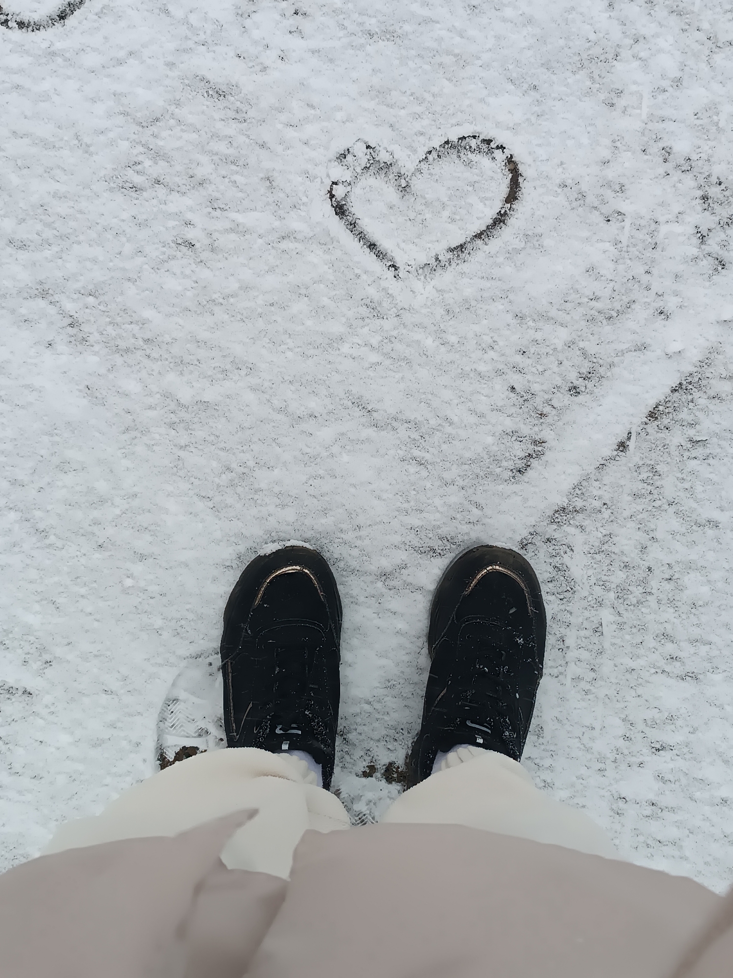 The image shows a first-person view of a person standing in snow. Two black sneakers are visible on the snow-covered ground. In front of the sneakers, a heart shape has been drawn in the snow. The location is outdoors in Unknown, Unknown.