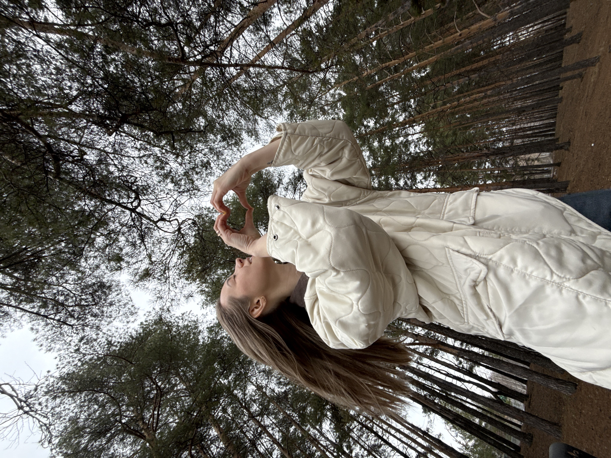 A woman is standing in a pine forest in Chaikovsky, Russia. She is wearing a quilted white jacket. She is holding her hands up to form a heart shape. The trees are tall and thin, and the ground is covered in pine needles. The sky is overcast.